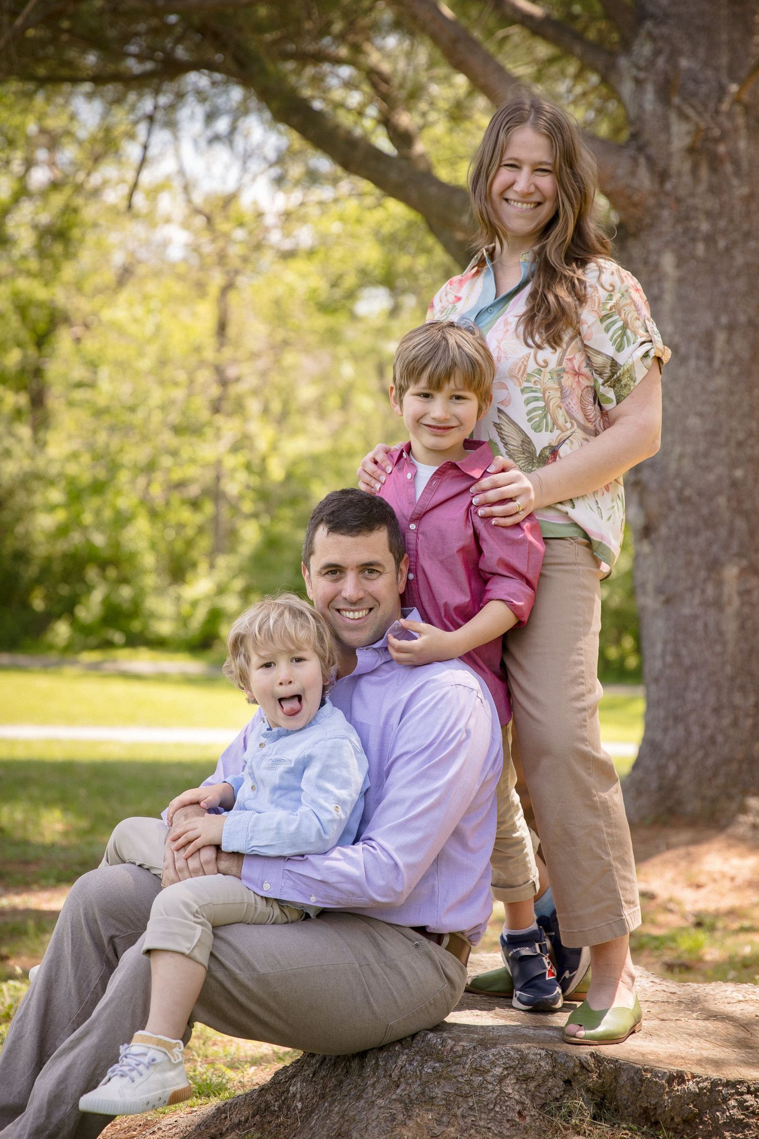 A smiling family of four poses outdoors under a large tree in a sunny park setting.