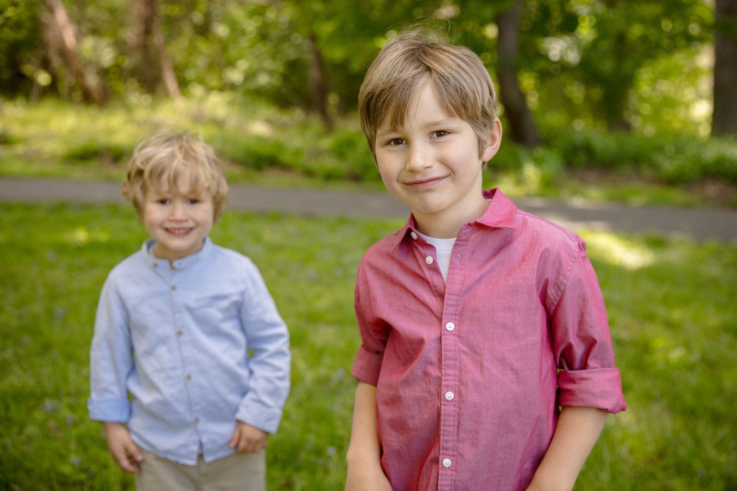 Two young boys smiling outdoors in a lush green park, wearing button-up shirts in blue and red.