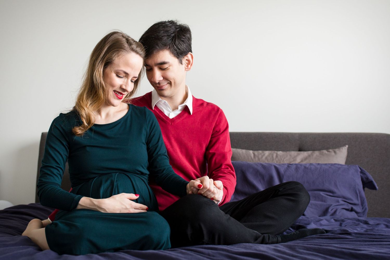 Pregnant woman in green dress sitting with partner in red sweater on bed, holding hands and smiling.