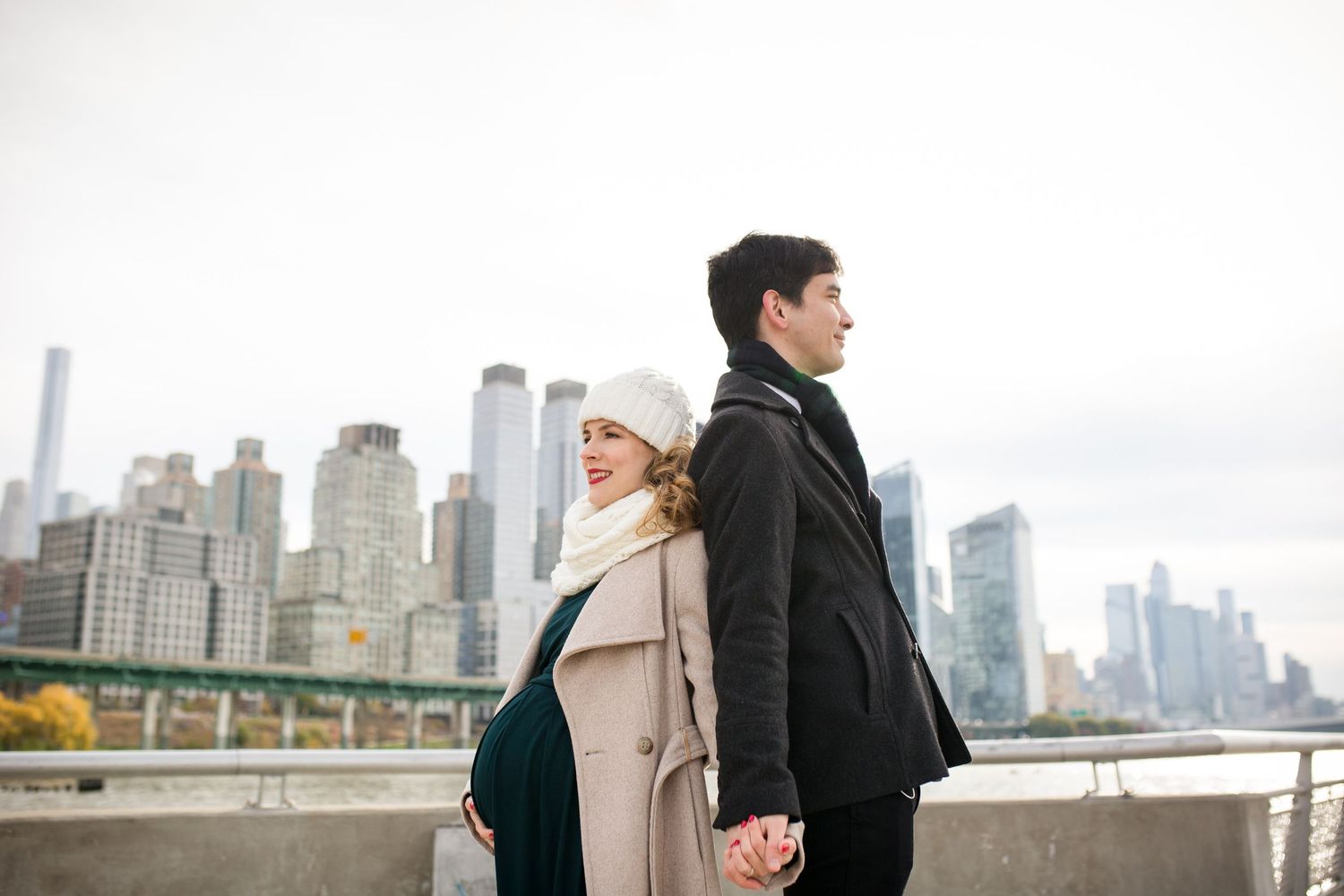 Pregnant couple holding hands back-to-back with NYC skyline backdrop on a cloudy day.