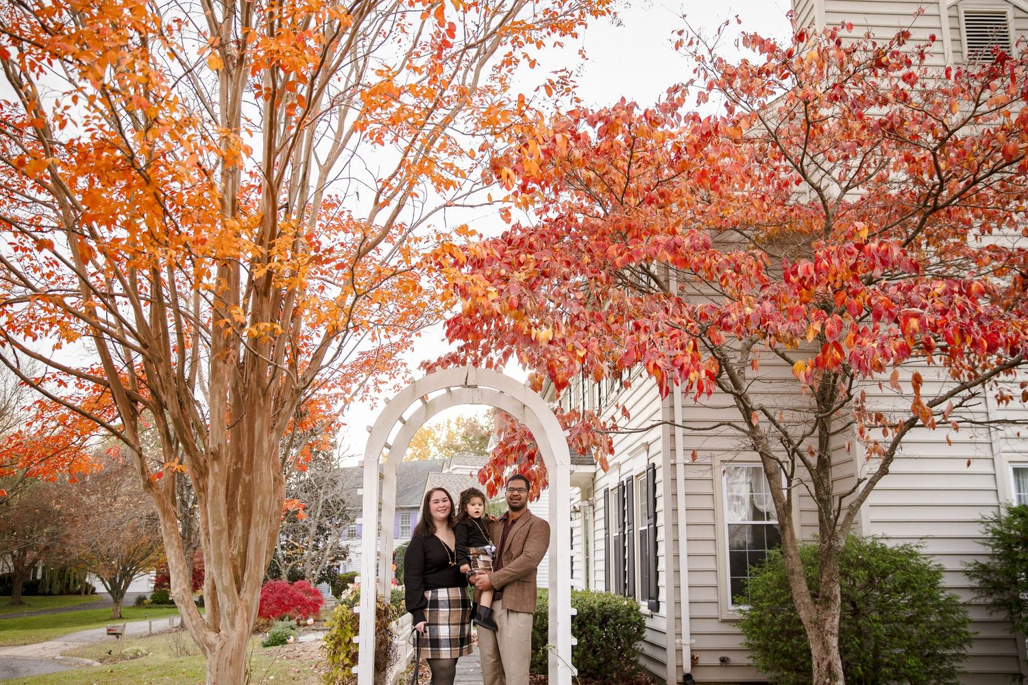 Family of three posing under white garden arch surrounded by vibrant red and orange autumn trees near a white house.