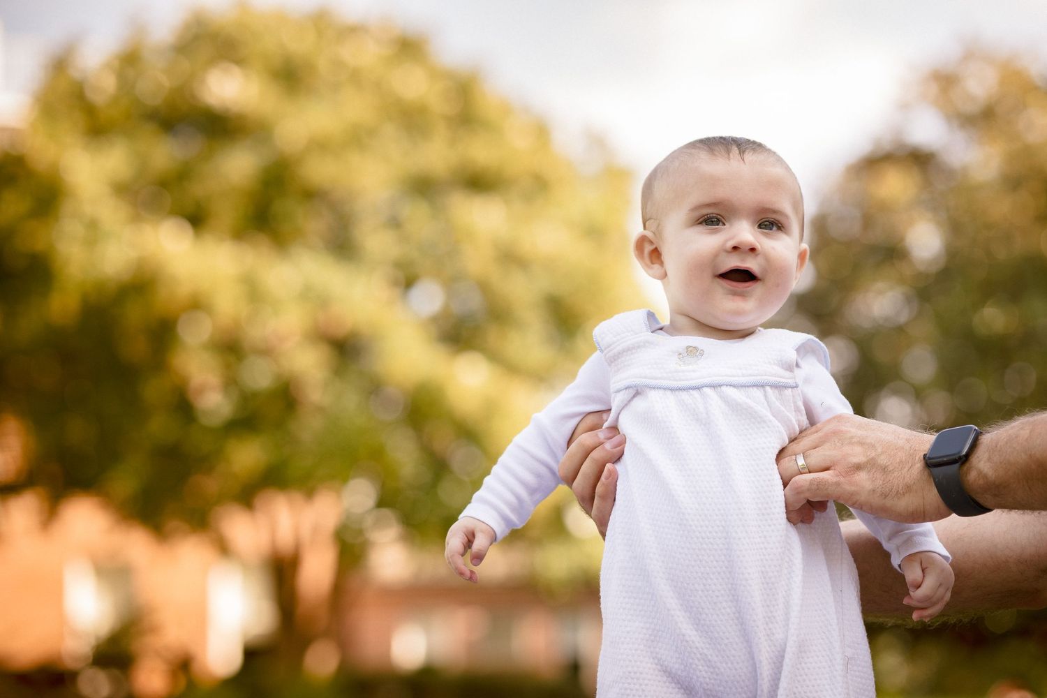 An adorable smiling baby in a white outfit being held up outdoors with colorful trees in background.