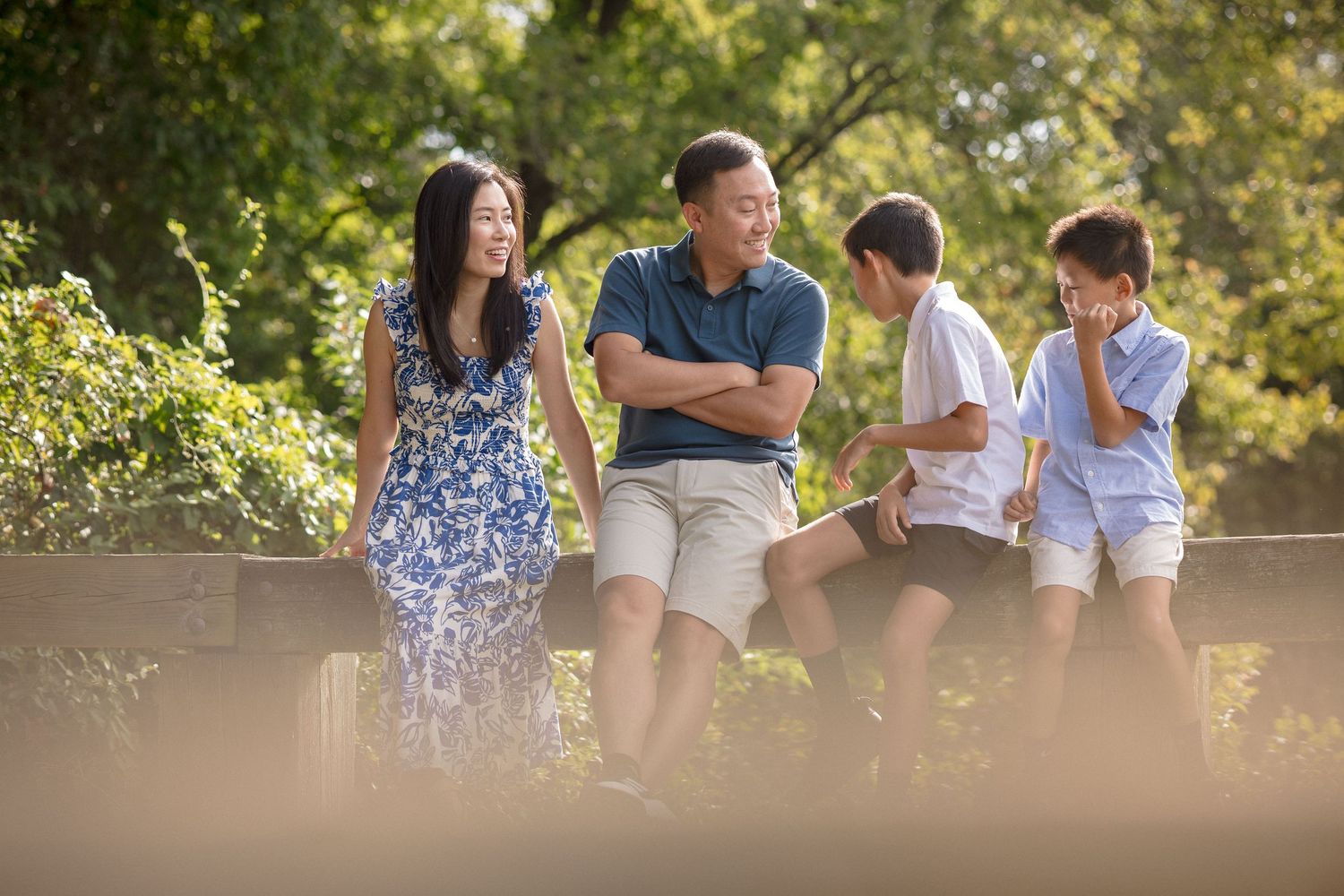 Asian family with two boys enjoying a sunny outdoor gathering, smiling and relaxing together near a wooden fence.