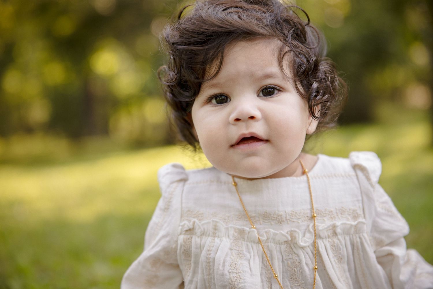 Adorable baby girl with curly dark hair wearing a white lace dress and gold necklace outdoors.