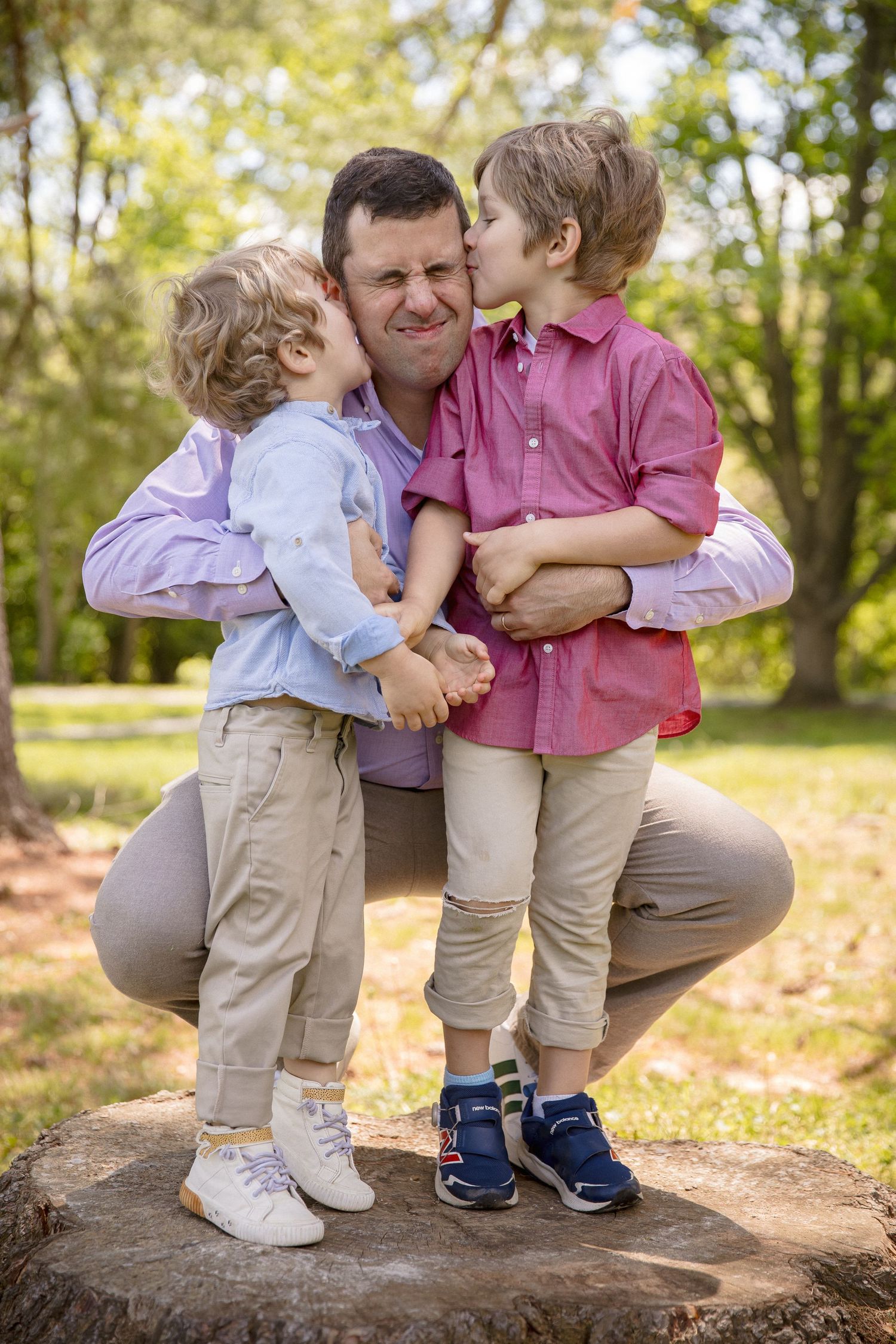 A father crouches on a tree stump in a park while his two young sons kiss him on both cheeks simultaneously.
