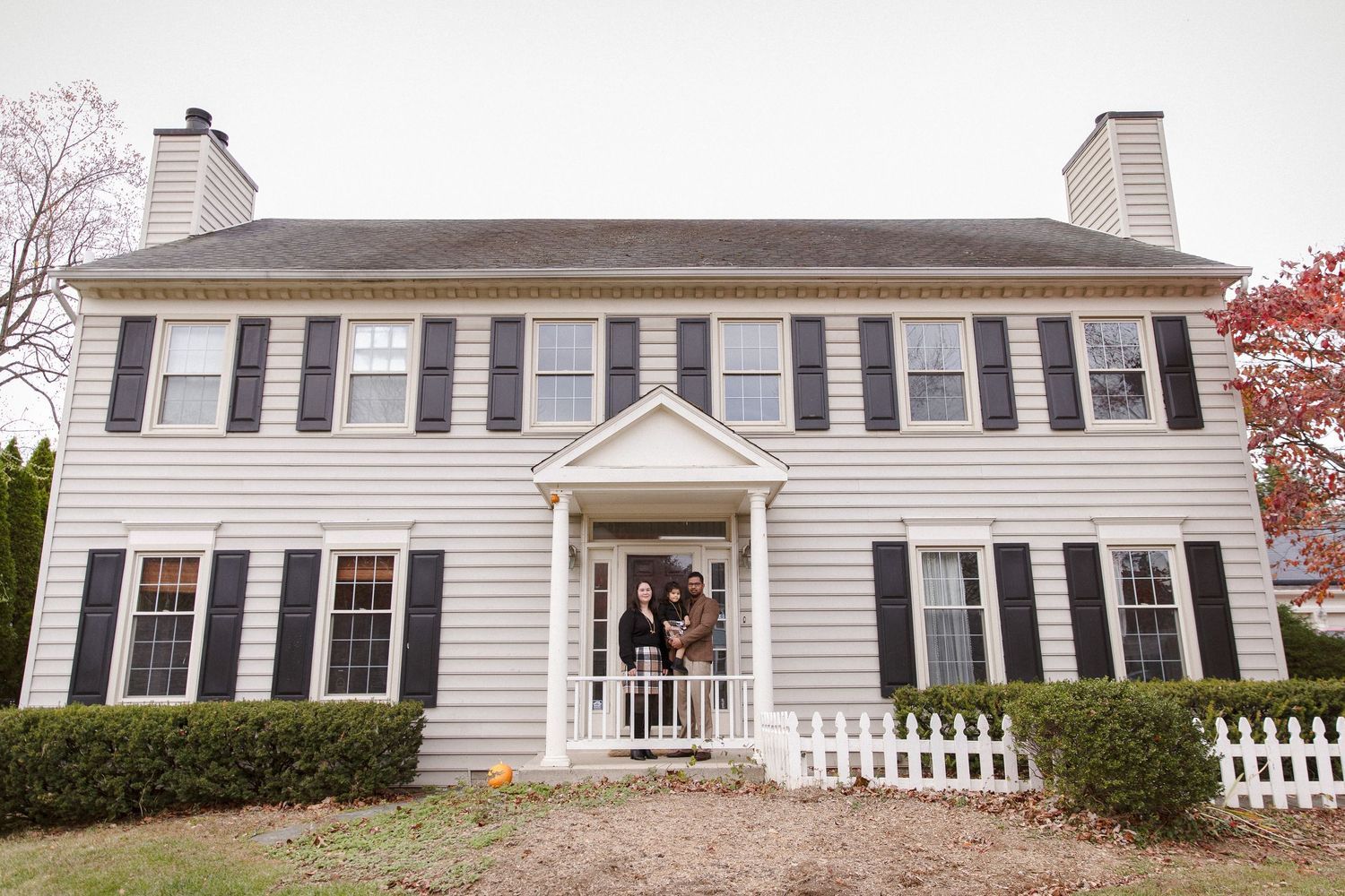 Family posing at the front door of a white colonial-style home with black shutters during autumn.