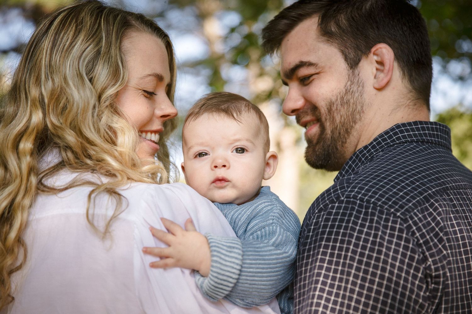 Young couple smiles lovingly at their infant baby outdoors among green trees.