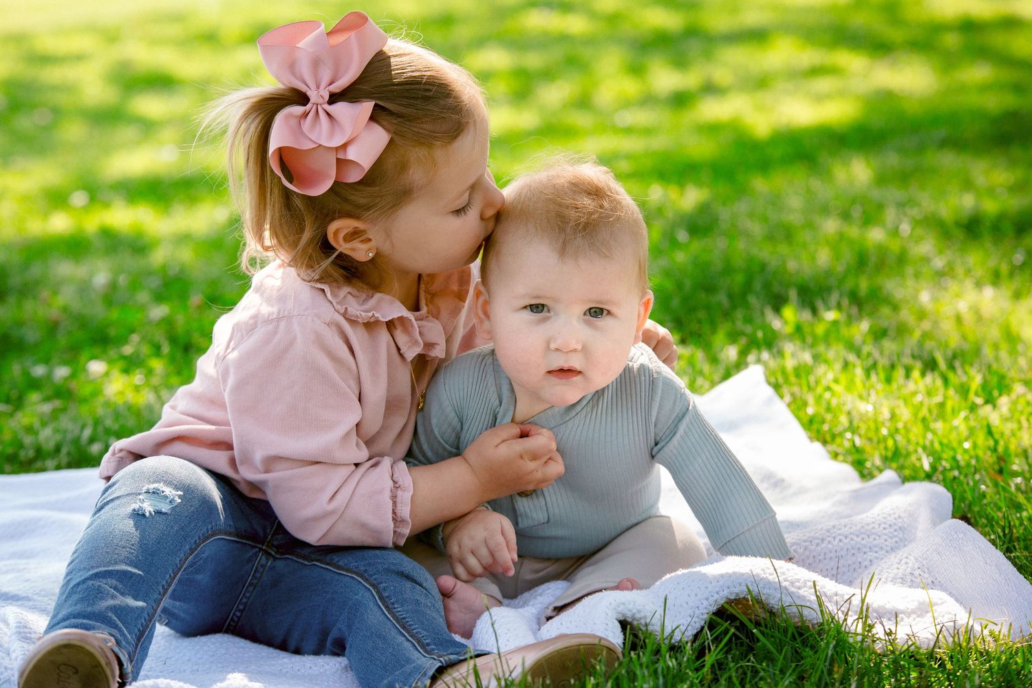 Young girl with pink bow kissing baby sibling on cheek while sitting on blanket in green grass.