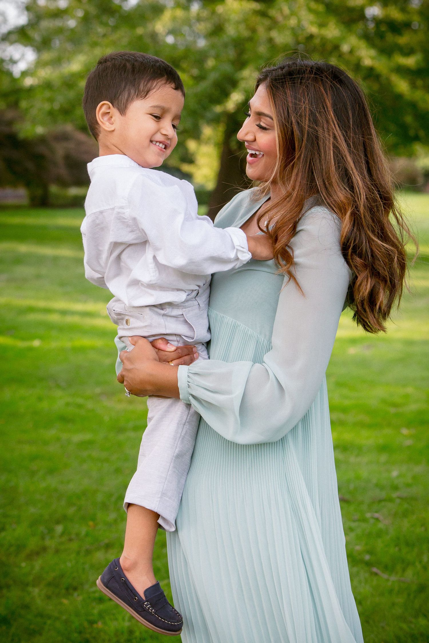 Smiling pregnant woman in mint dress holding laughing toddler boy in white shirt at a green park.