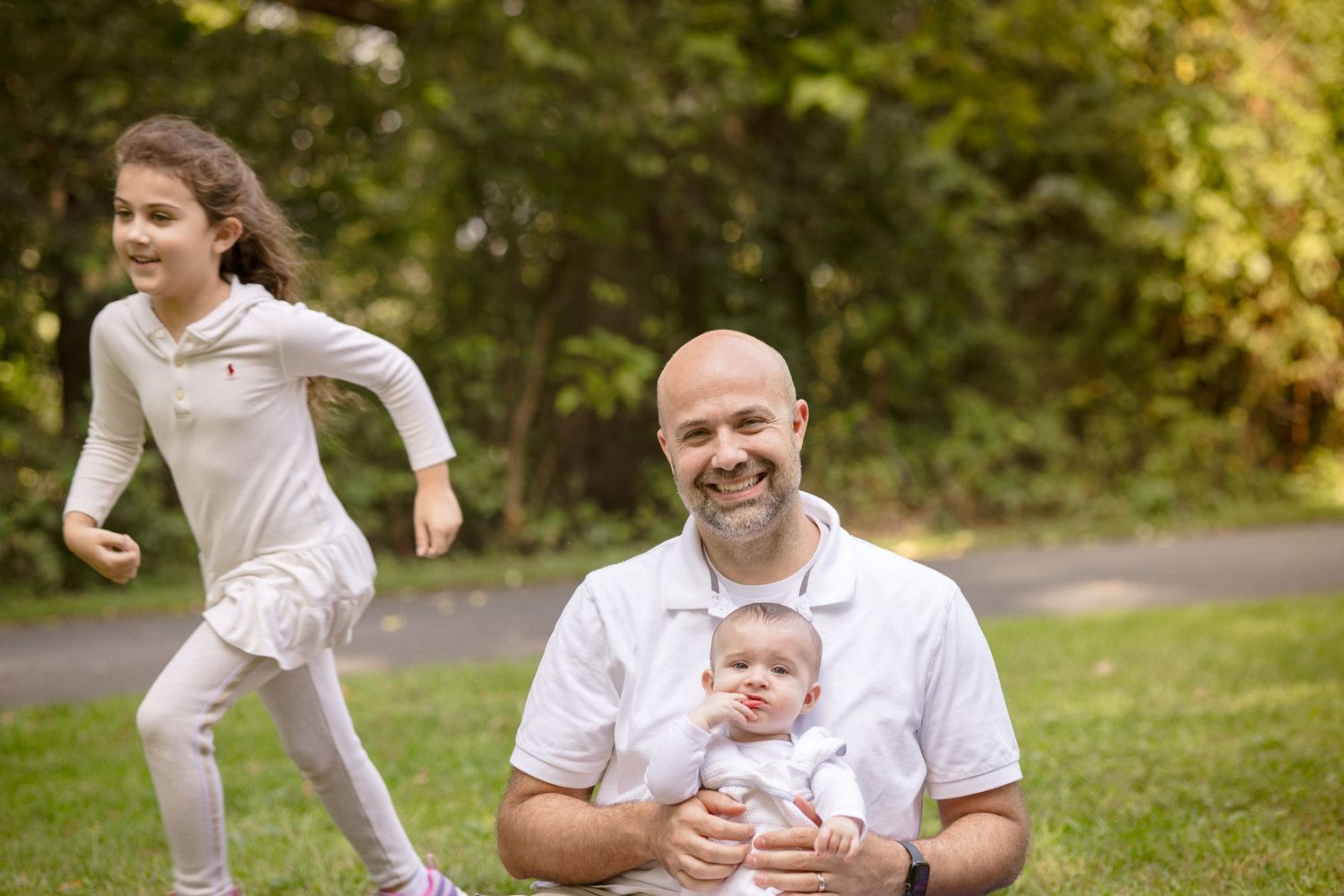 A bald father sits on grass holding a baby while his young daughter runs playfully behind him.