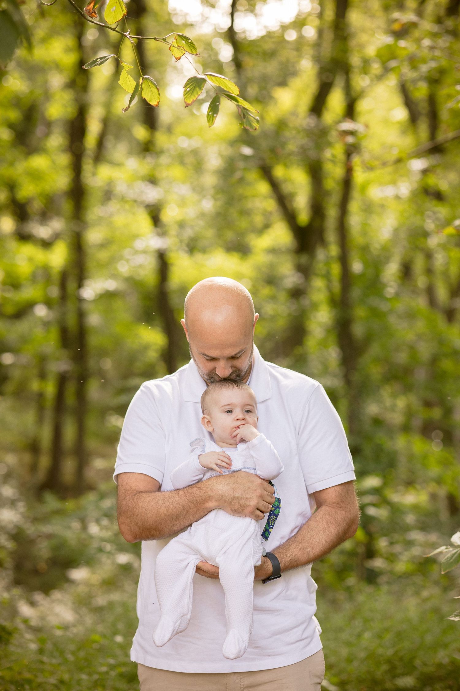 Bald father with beard tenderly holds baby in white onesie outdoors in lush green forest setting.