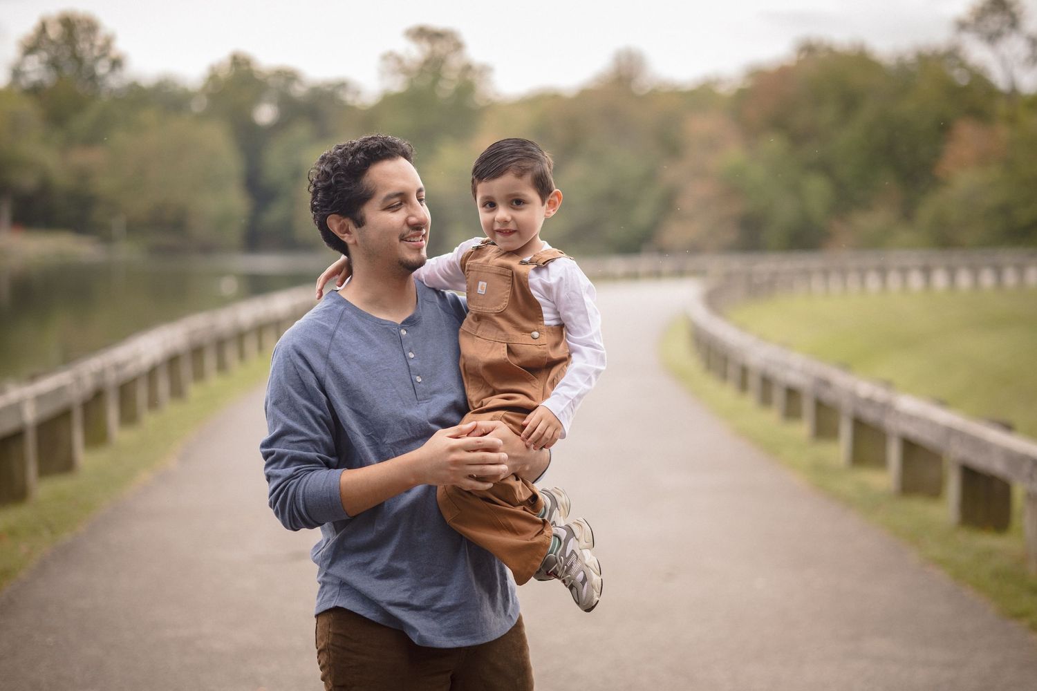 A father joyfully carries his young son on a scenic park path surrounded by autumn trees near a lake.