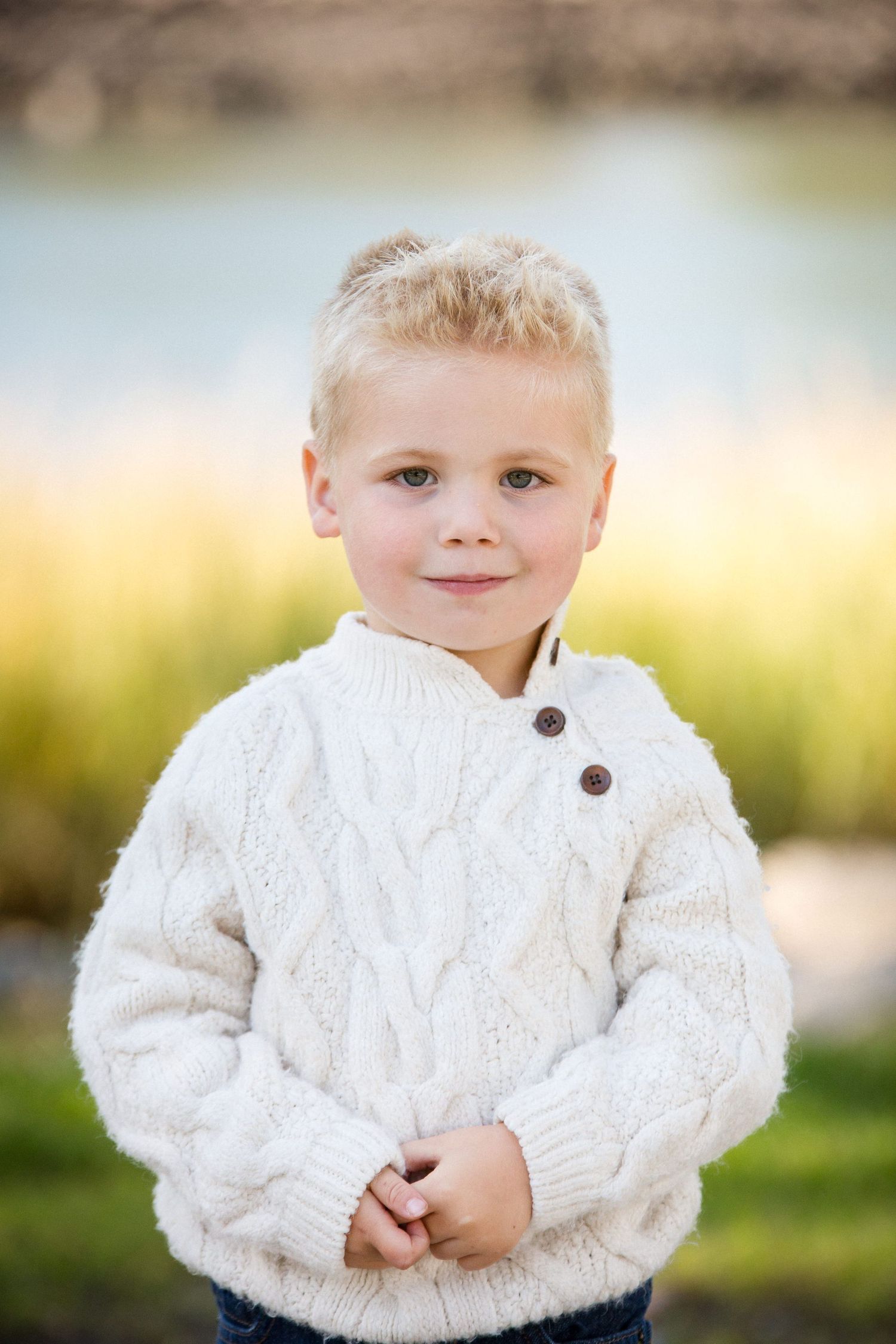Smiling young boy with blonde hair wearing a white cable-knit sweater outdoors near a pond.