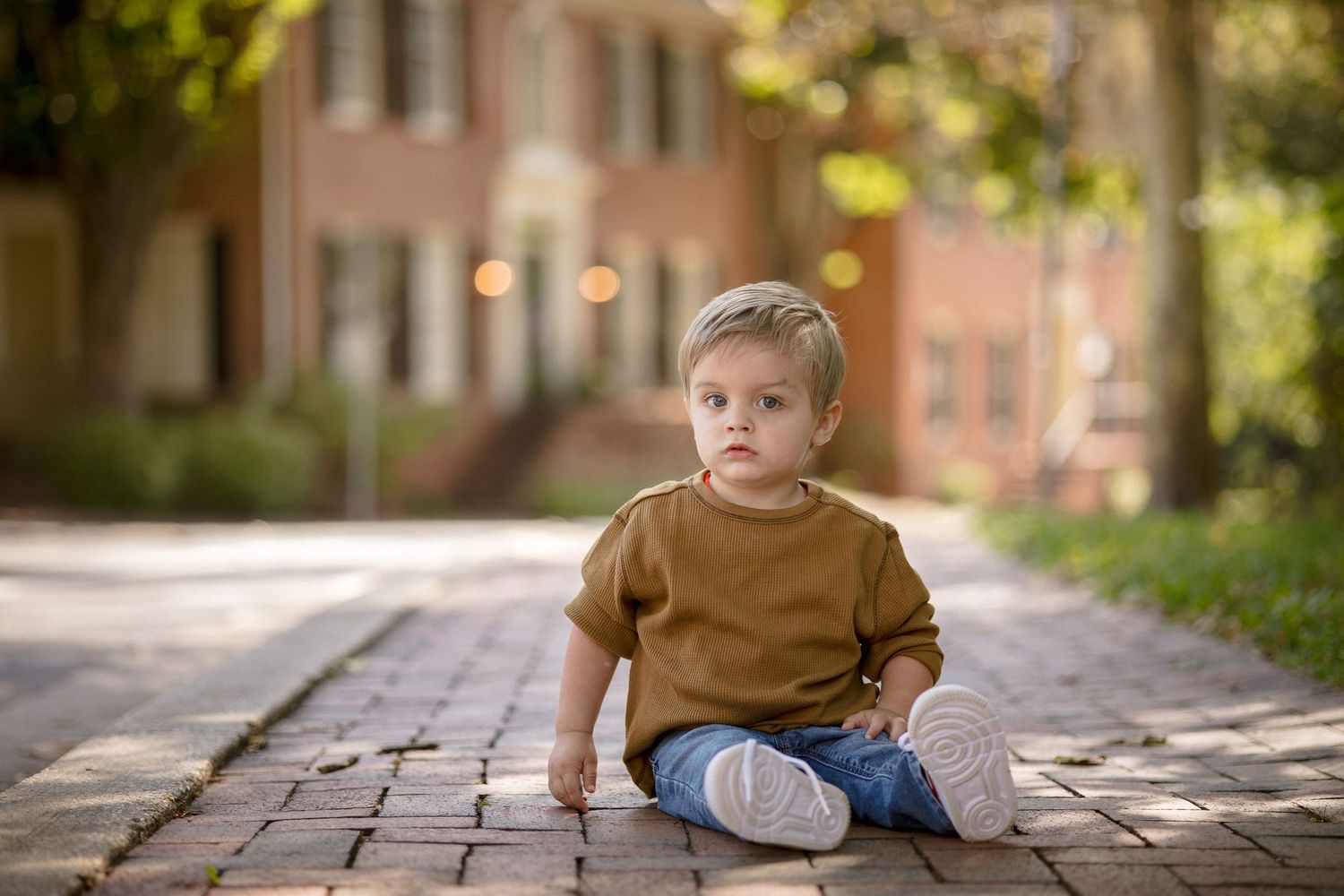Toddler boy in brown sweater and jeans sitting on brick path outdoors with blurred building background.