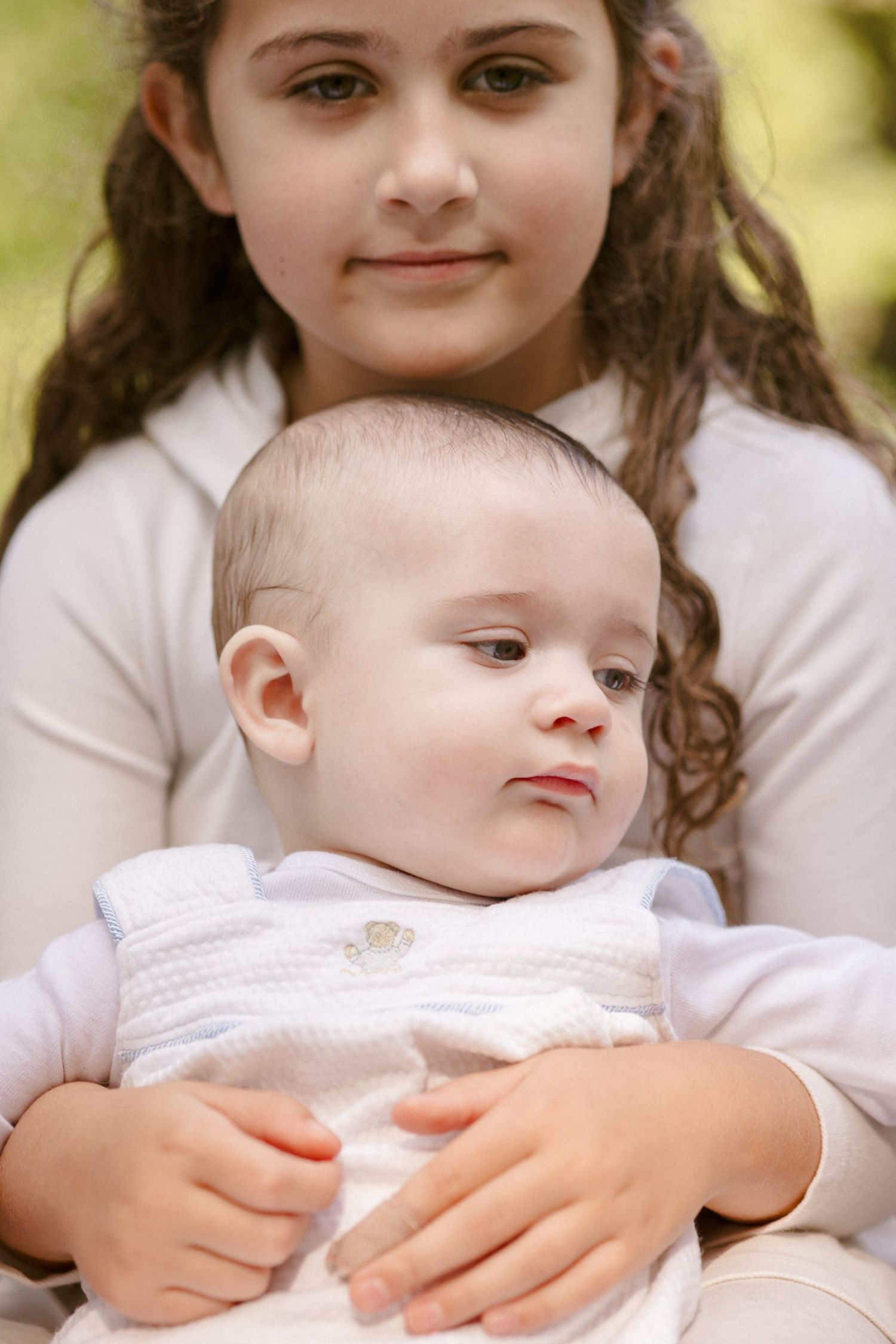 Older girl with curly hair gently holding a baby in white outfit outdoors in a green park setting.