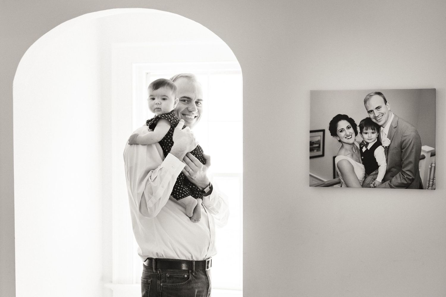 Black and white photo of a father holding his toddler near a framed family portrait.