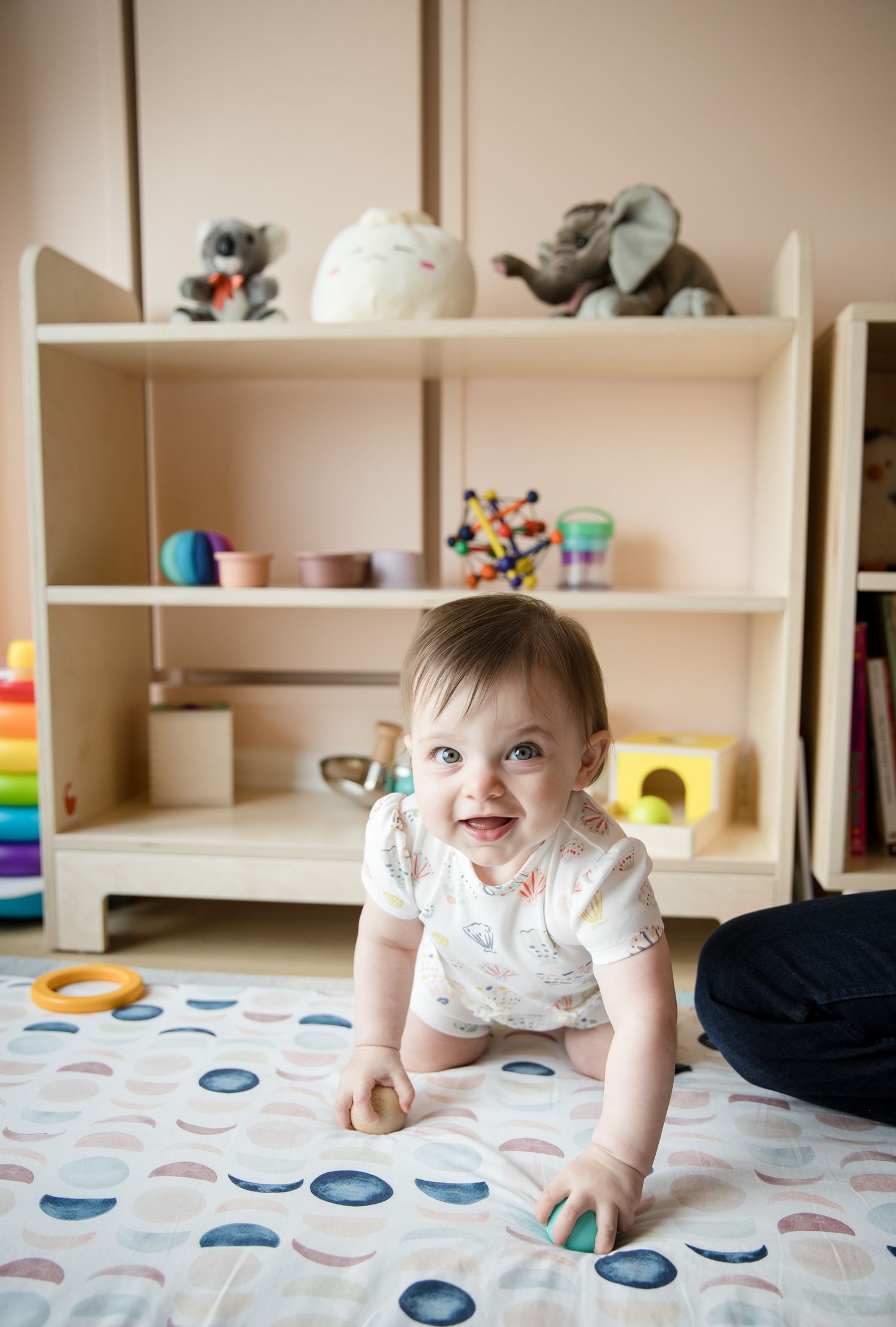 Smiling baby crawling on a patterned play mat in a cozy nursery with wooden shelves and colorful toys.