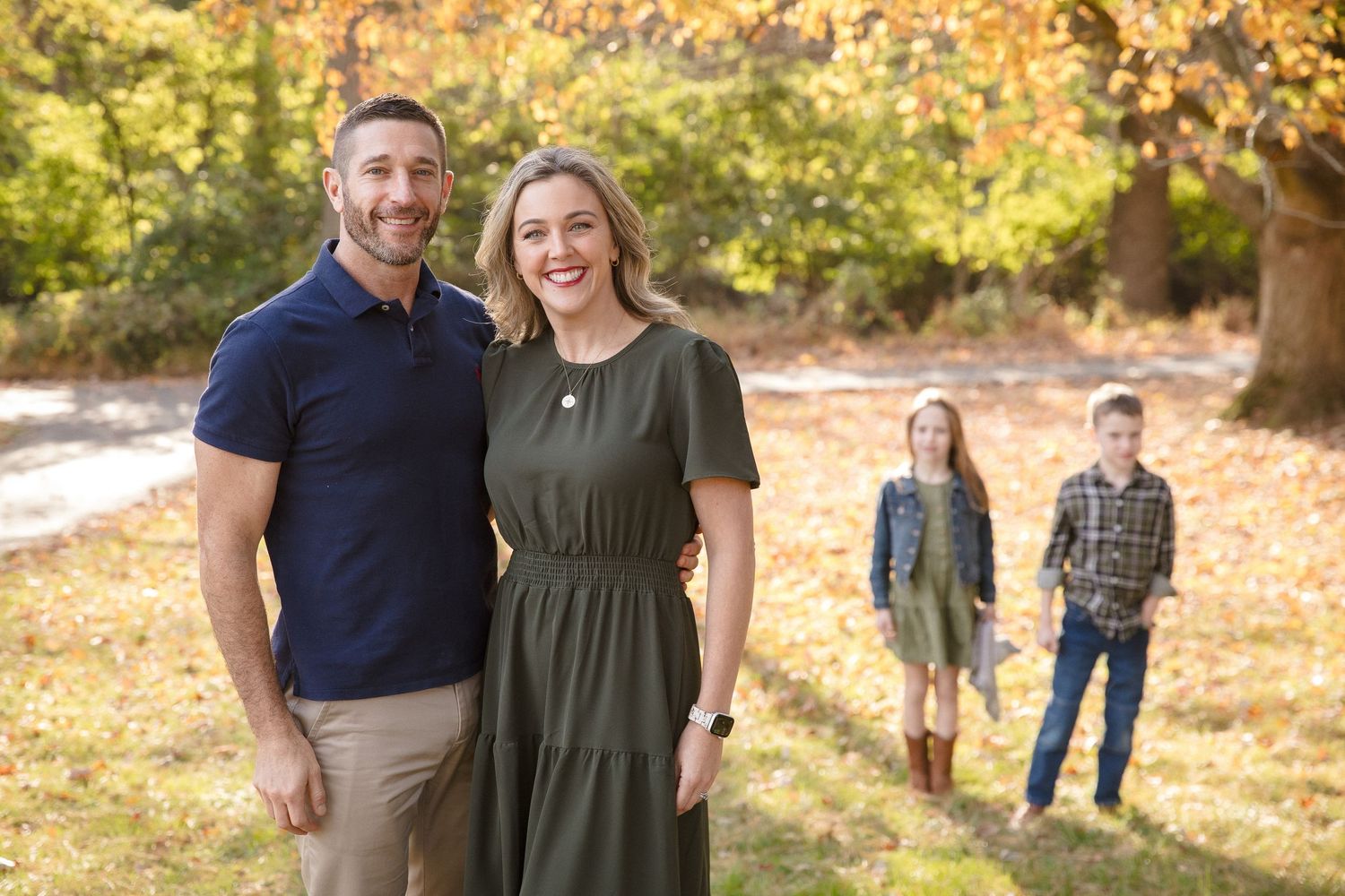 Smiling couple in autumn park with two children visible in the colorful fall foliage background.