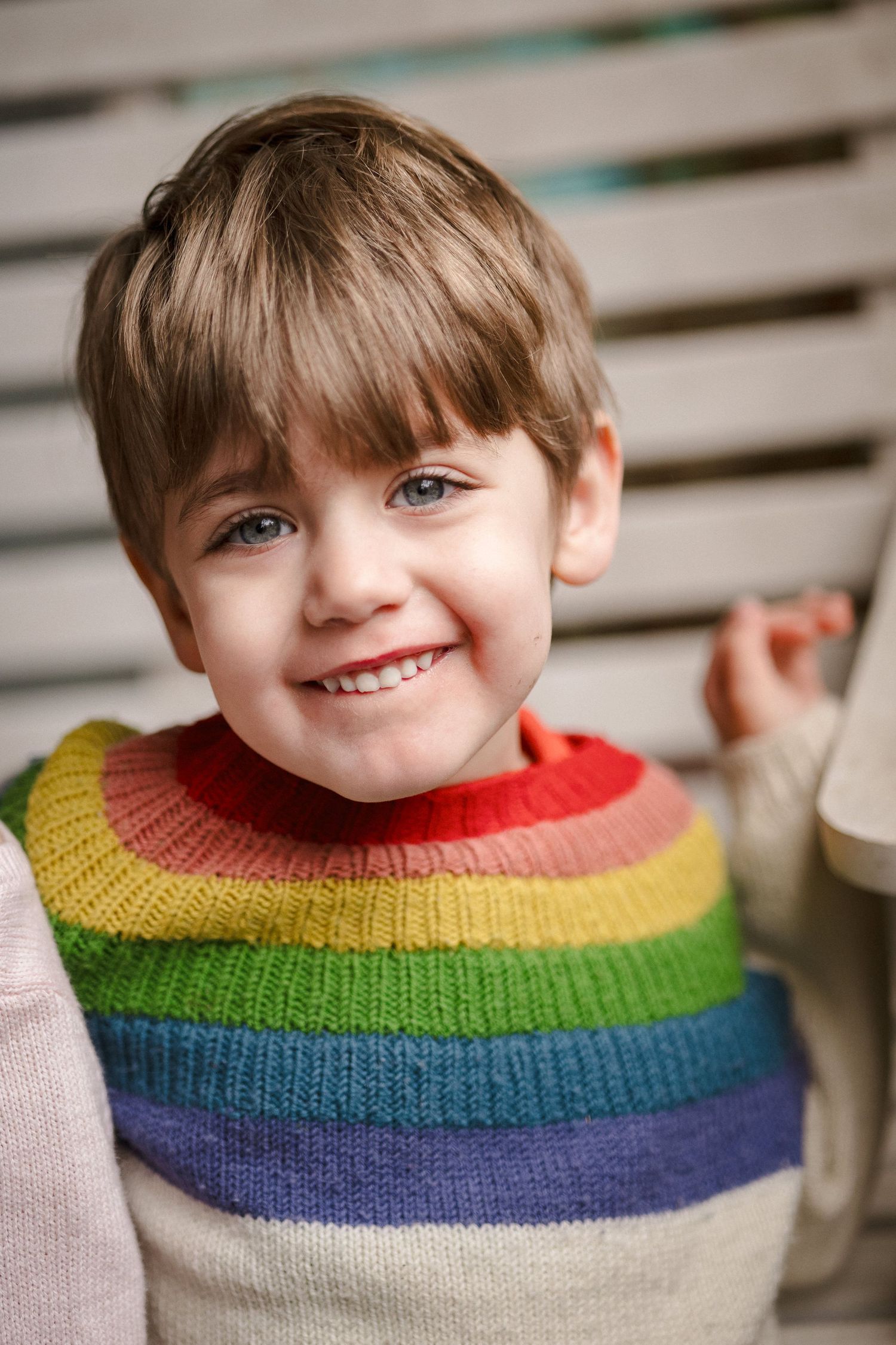 Cheerful young boy smiling brightly wearing a colorful rainbow striped knit sweater indoors.