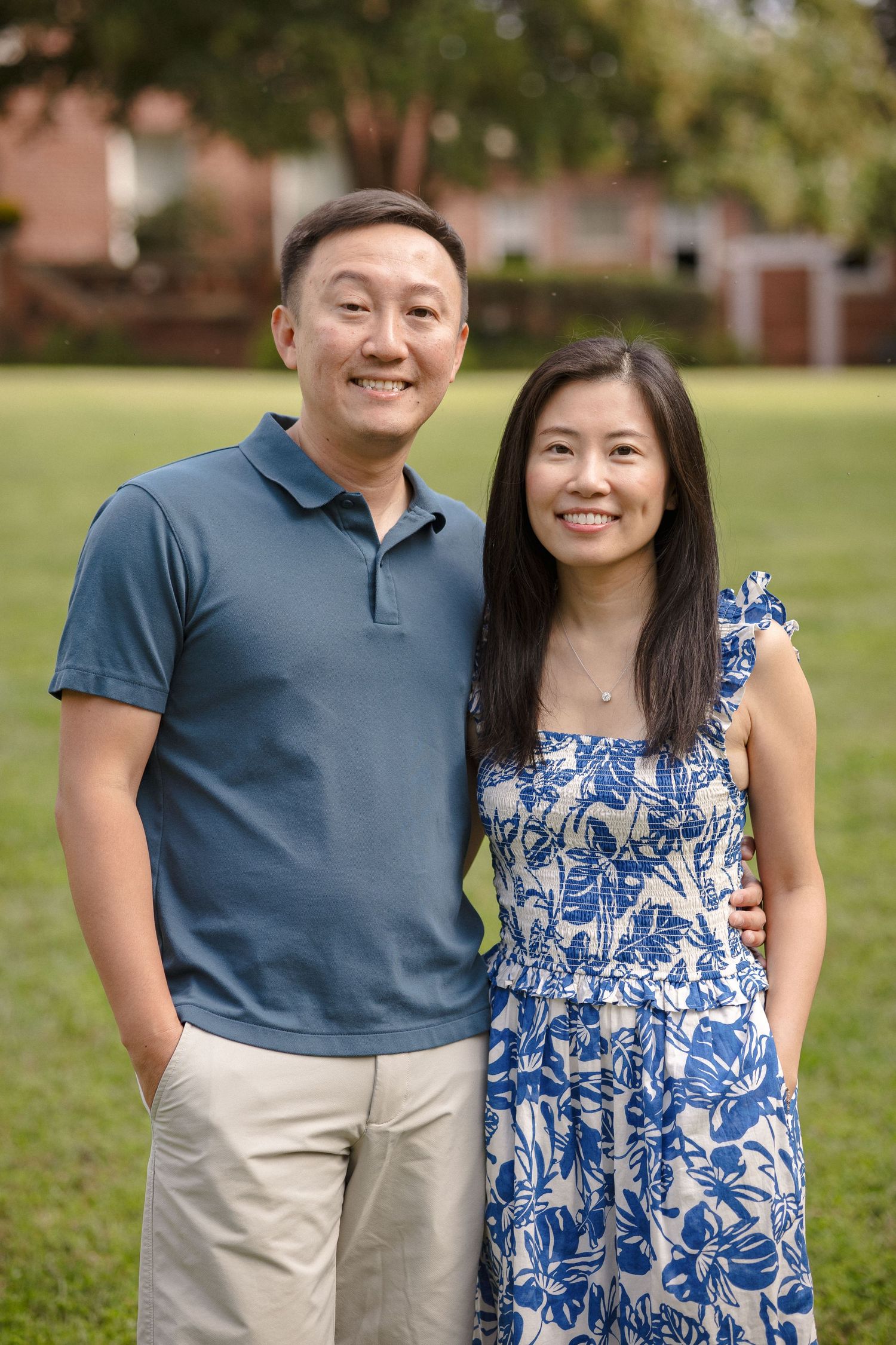 Smiling Asian couple posing outdoors on green grass, man in blue polo and woman in floral dress.