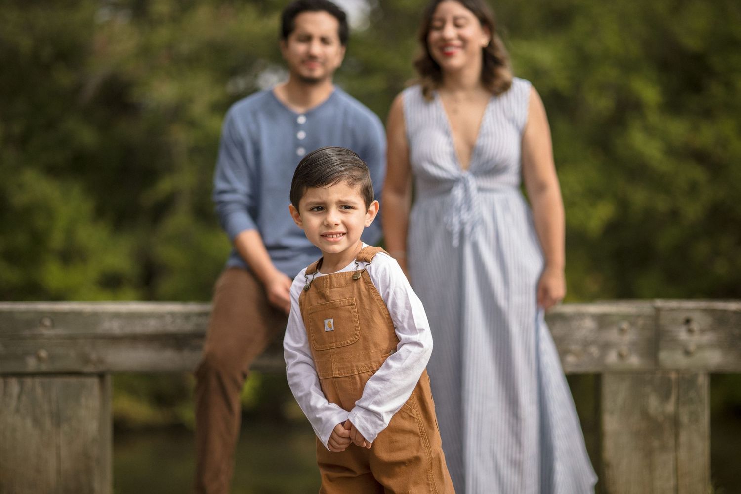 Young boy in brown overalls smiles at camera with parents standing on wooden bridge in soft bokeh background.