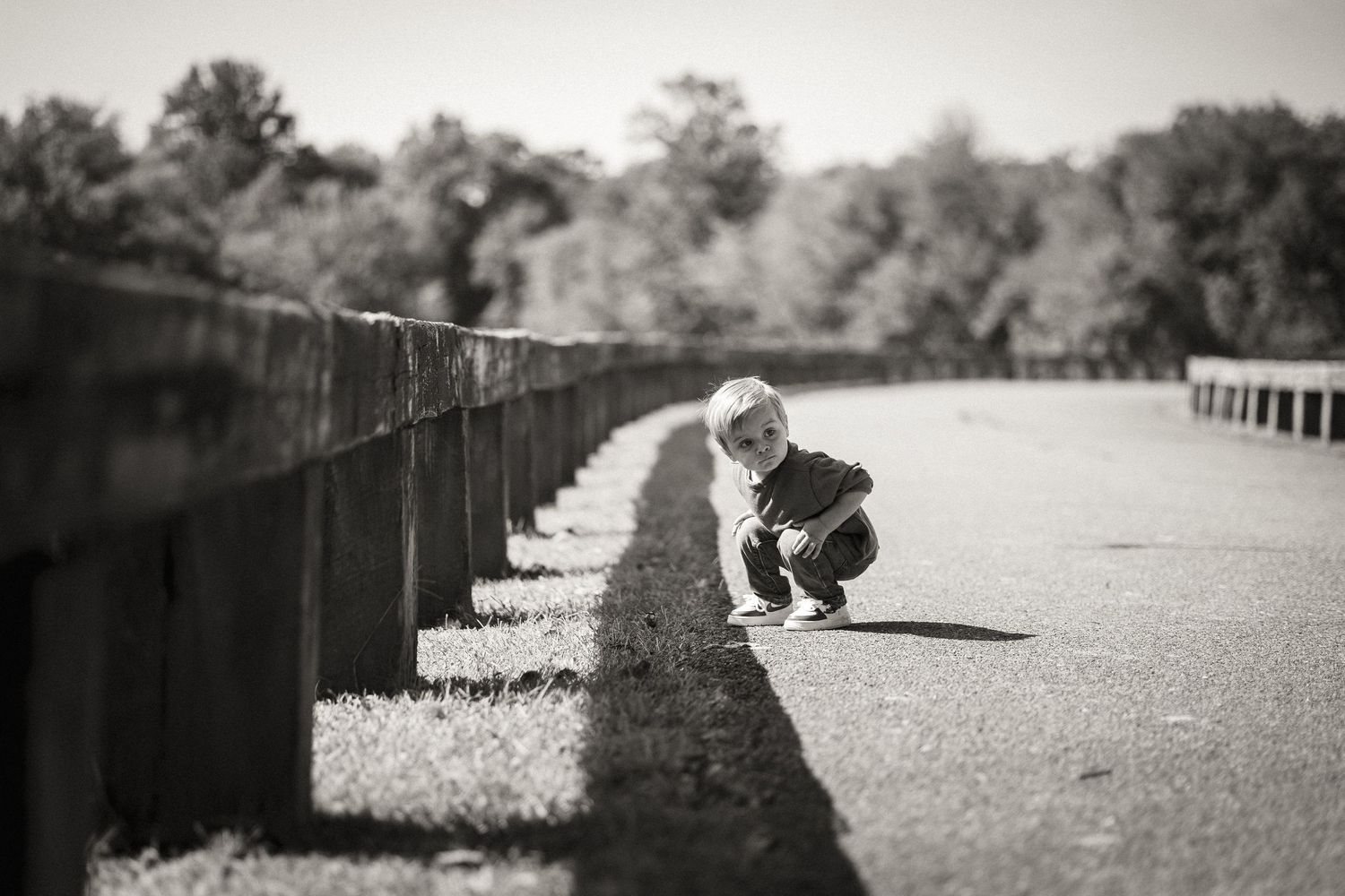 Black and white photo of a young child crouching on a road beside a concrete barrier with trees in the background.