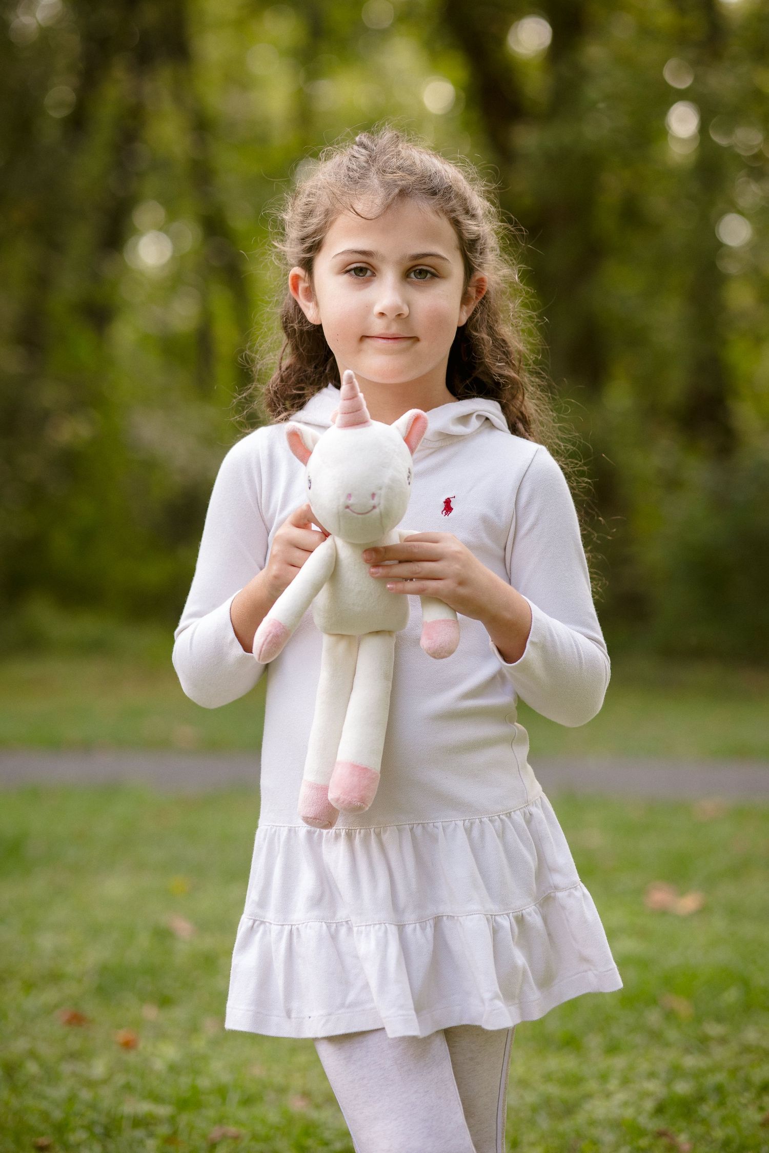 Young girl in white dress holding a unicorn stuffed animal toy while standing outdoors in a green park.