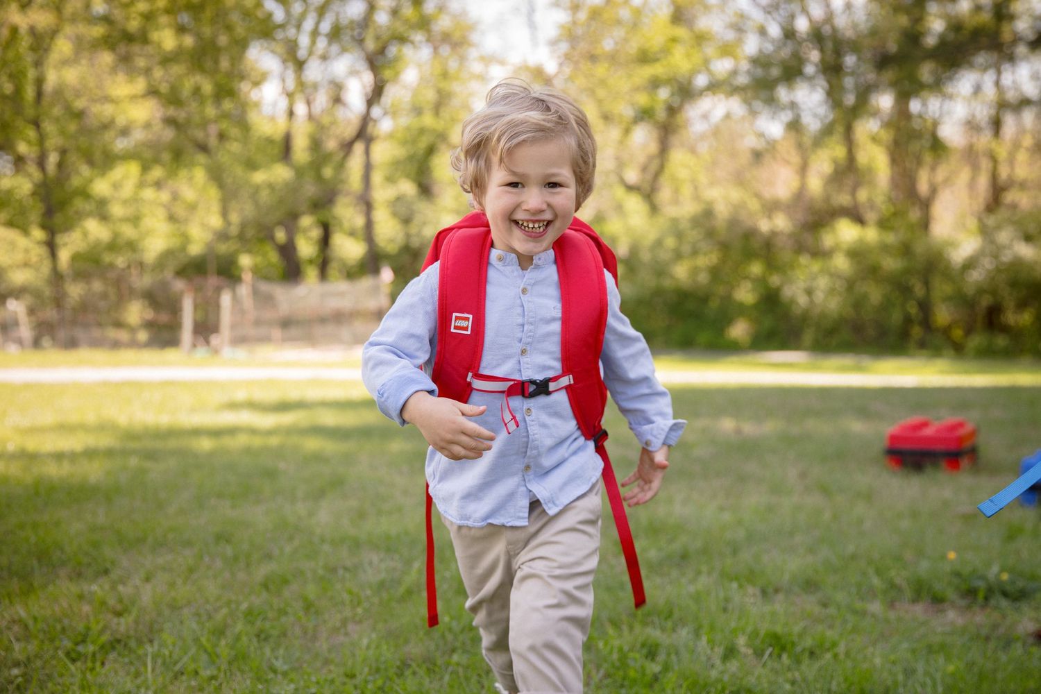 Young boy wearing a red backpack smiling and running on green grass in a sunny outdoor park setting.