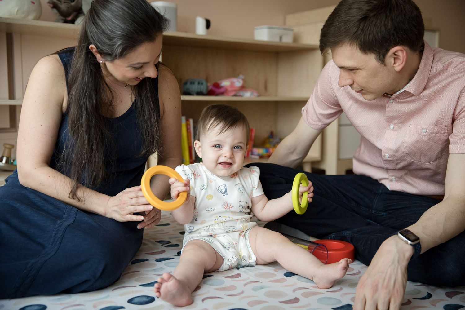 Happy baby sits between smiling parents playing with colorful ring toys on a blanket in a bright nursery room.