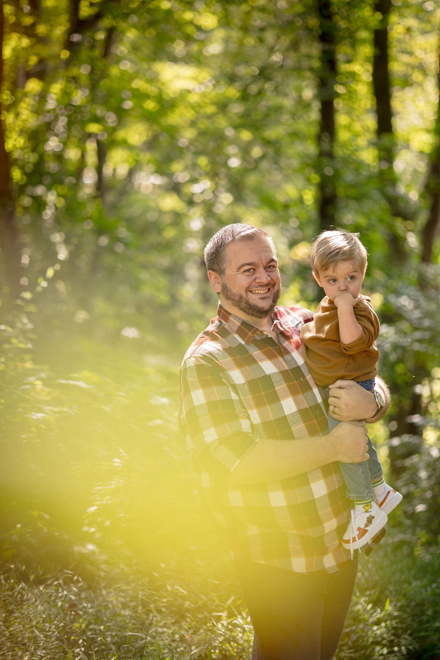 A smiling father holds his young toddler son in a sunlit green forest, both wearing casual warm-toned clothing.