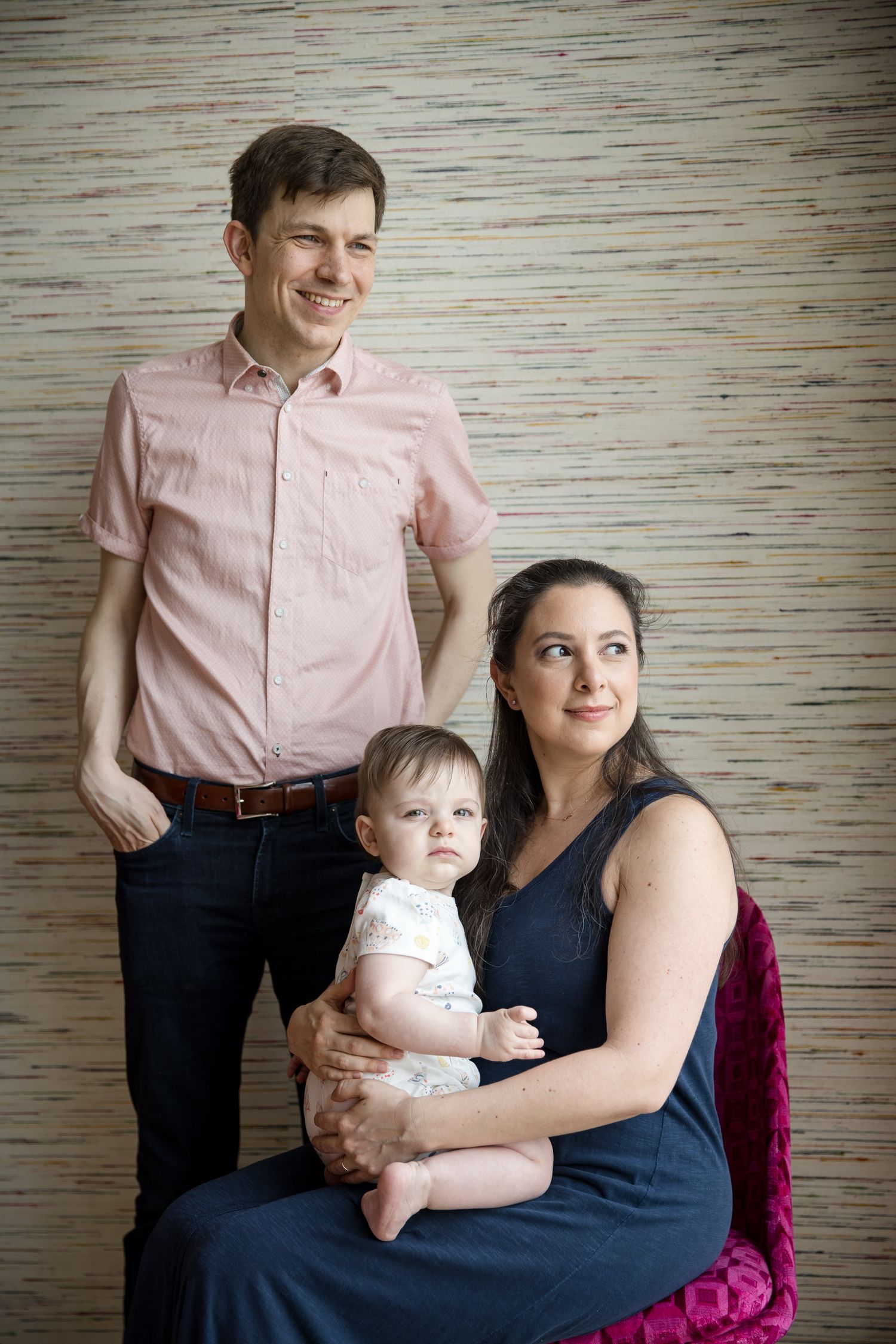 A smiling family portrait with a father standing, mother seated holding a baby, posed against a textured wall backdrop.
