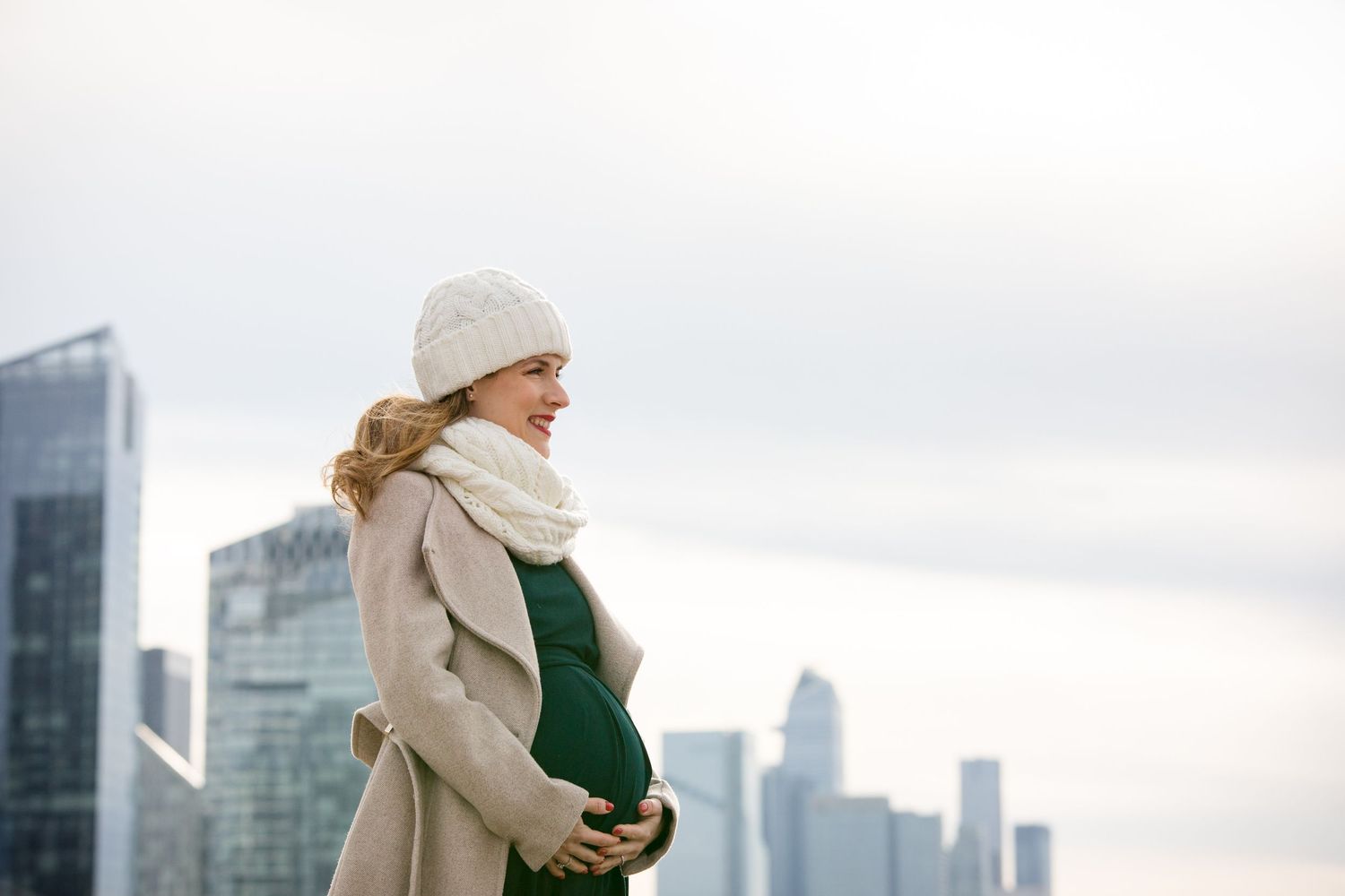 Smiling pregnant woman in winter coat and knit hat cradling baby bump against city skyline backdrop.