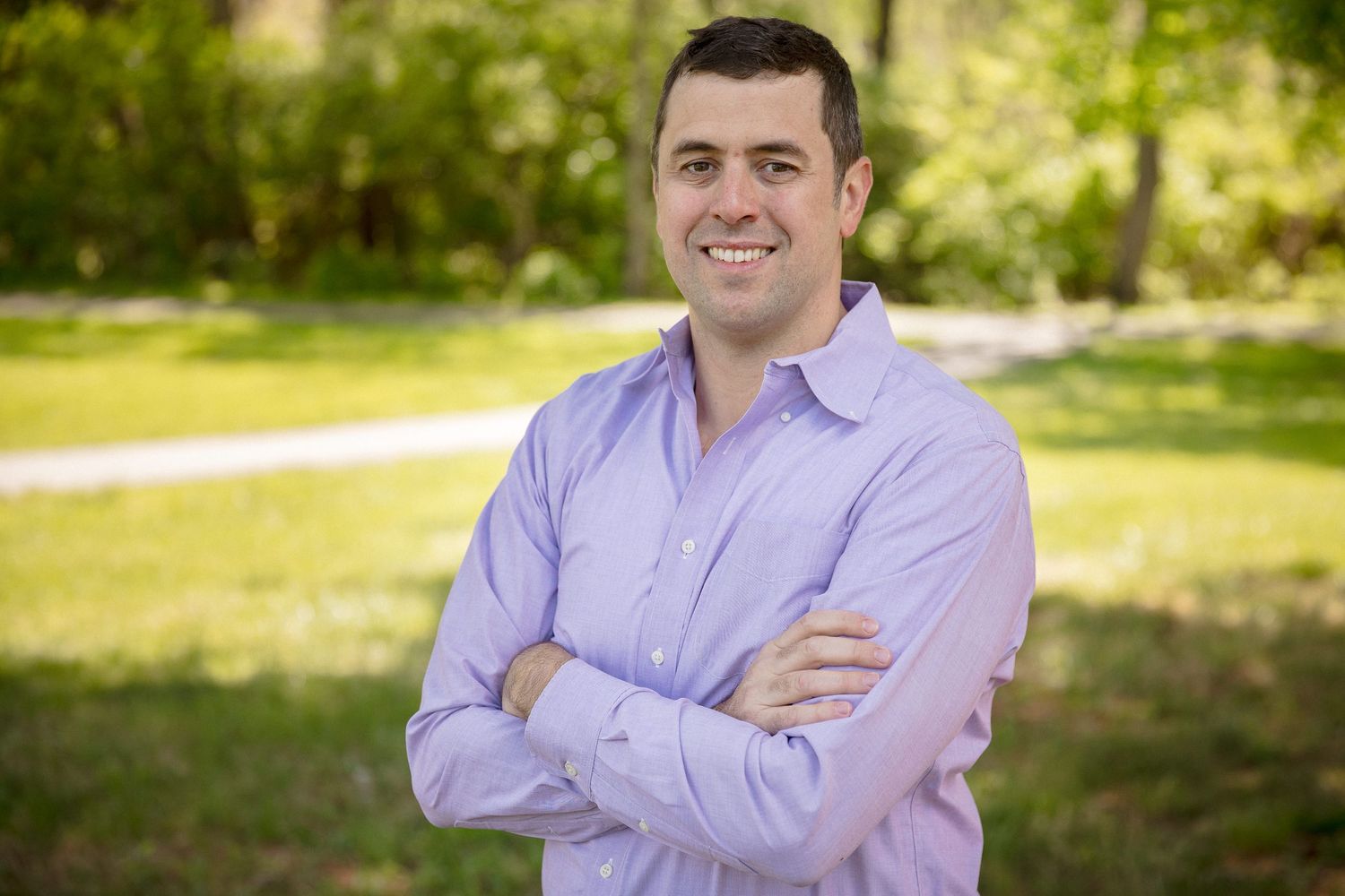 Smiling man in lavender shirt standing outdoors with arms crossed in a lush green park setting.