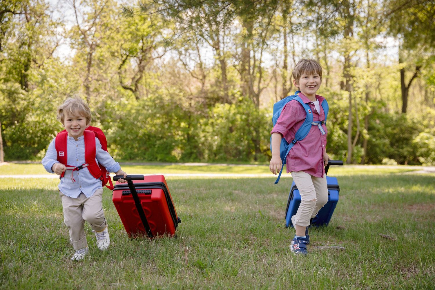 Two young children running in a park with backpacks and luggage, smiling on a sunny day.