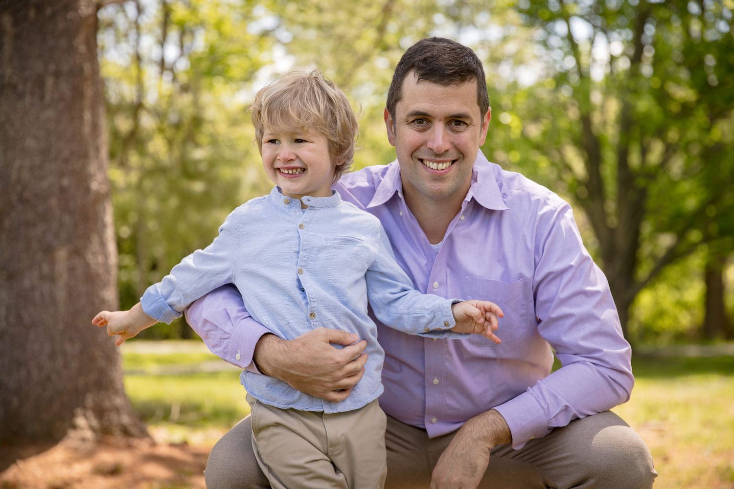 A smiling father crouches beside his young blond son outdoors in a lush green park setting.