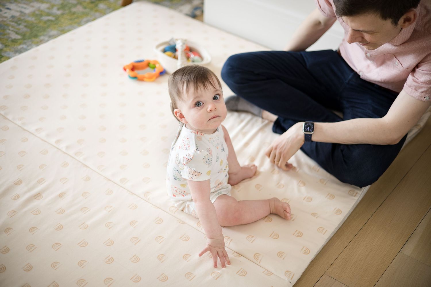 Baby sitting on a padded play mat with an adult nearby, surrounded by colorful toys in a bright room.
