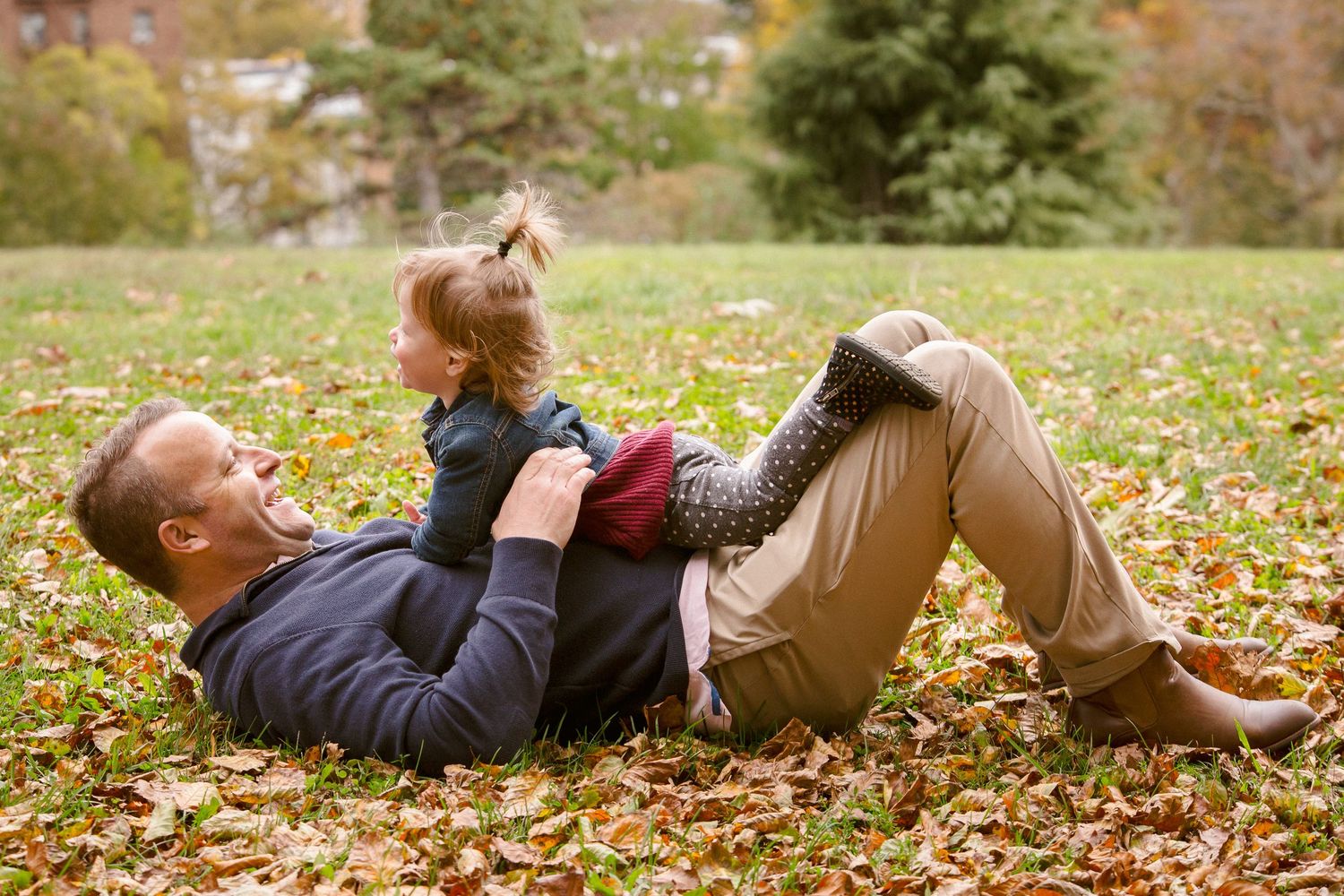 Father laughing while playing with young daughter on fallen autumn leaves in park.
