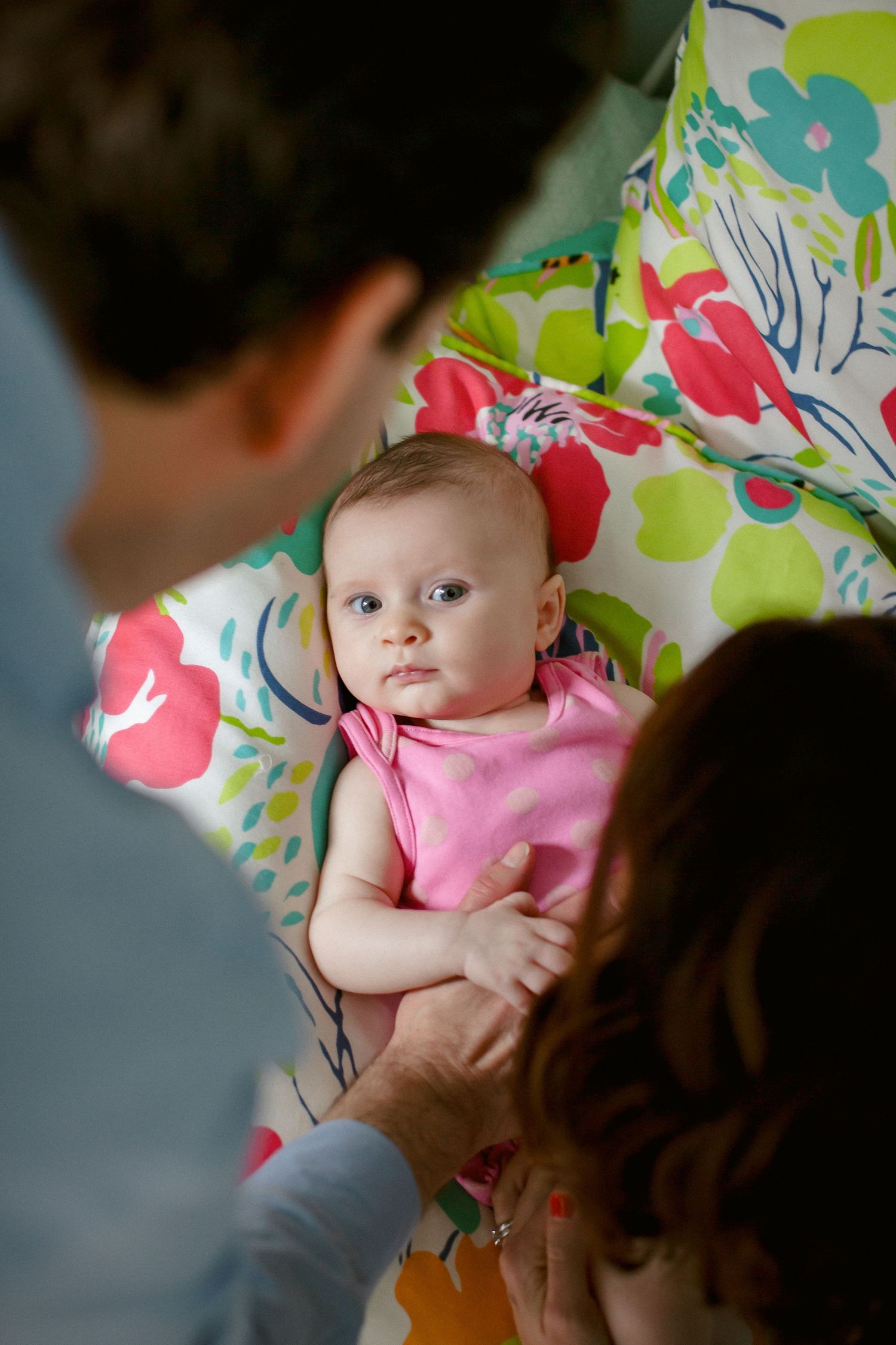 Parents lovingly attend to their infant baby dressed in pink on a colorful floral chair.
