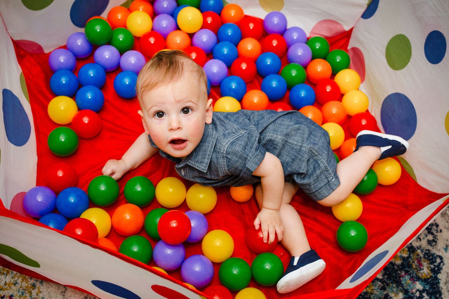 Baby boy lying in colorful ball pit smiling at camera.