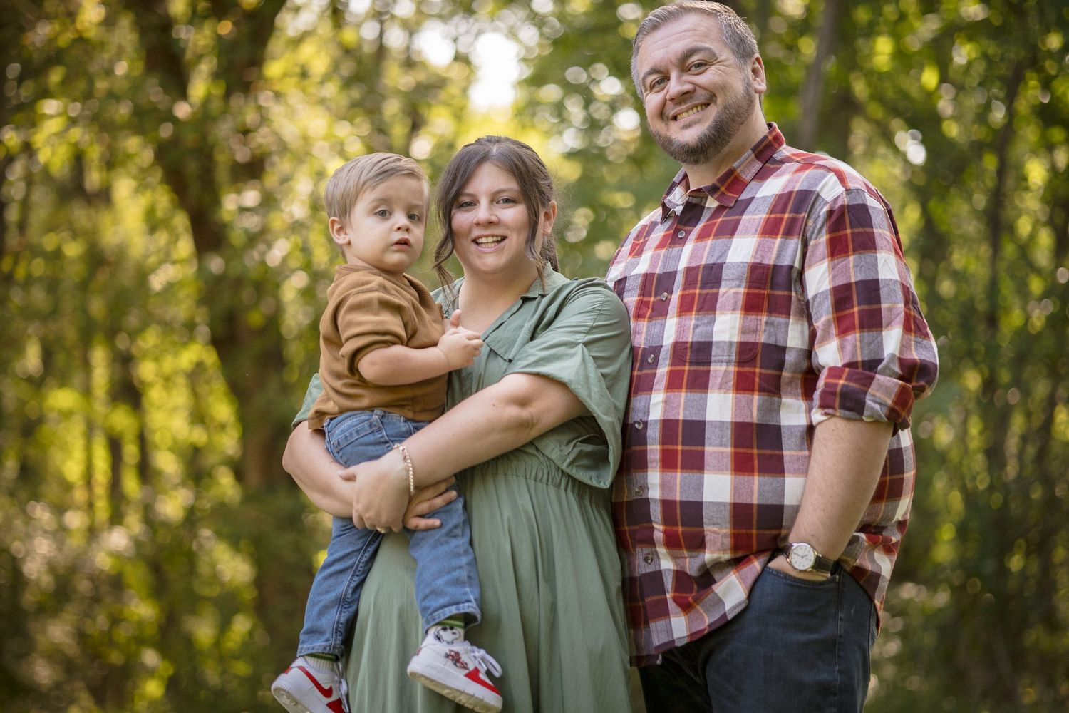 Happy family portrait with parents and toddler smiling outdoors in lush green forest setting.