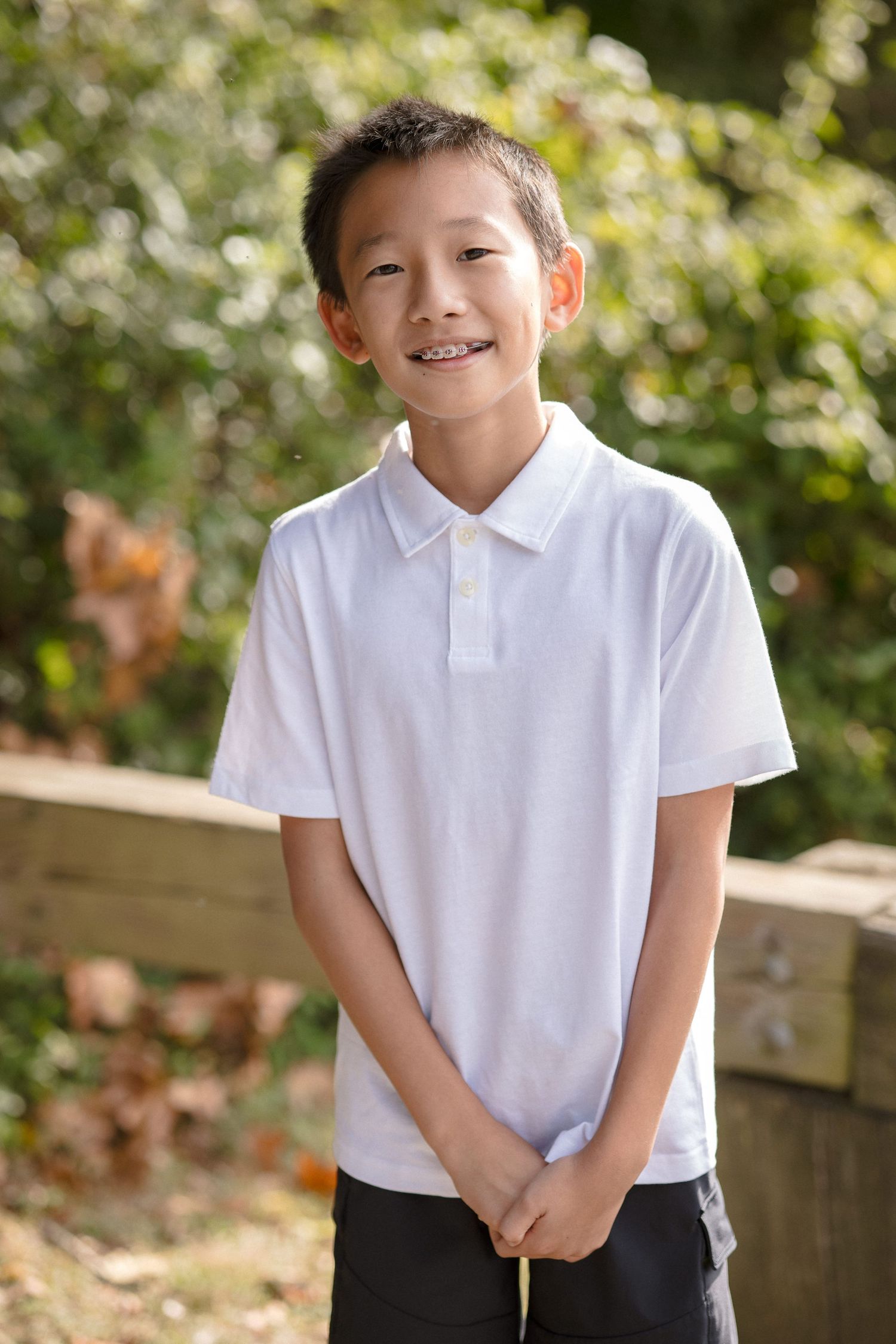 Smiling Asian boy in white polo shirt standing outdoors in a garden setting with green foliage background.