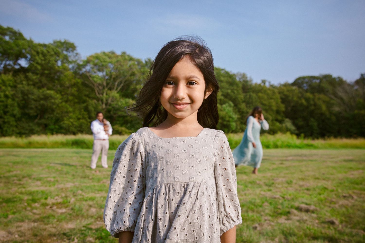 Young girl in white dress smiling in green field with family in background.