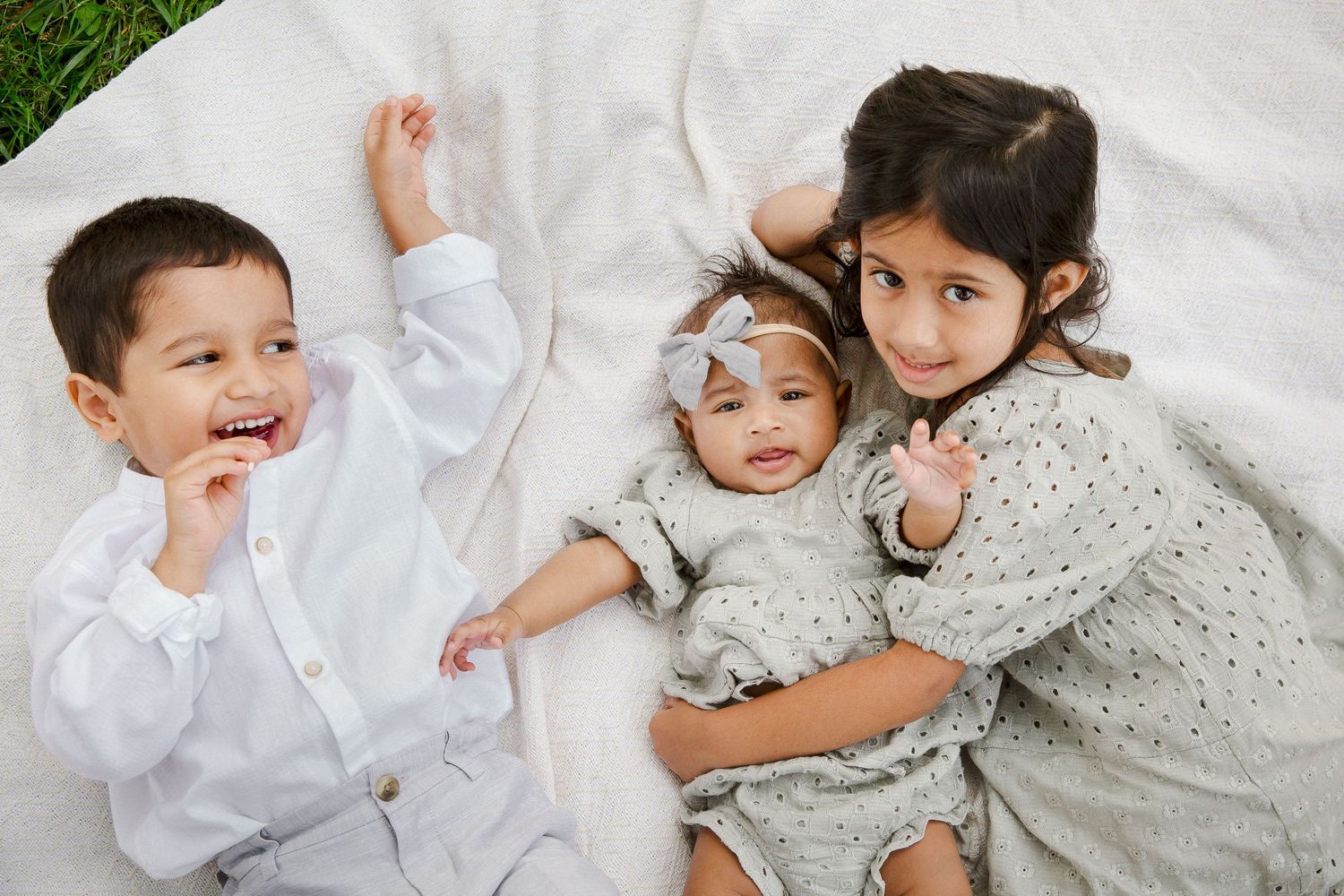 Three young siblings lying together on a white blanket outdoors, smiling at the camera.