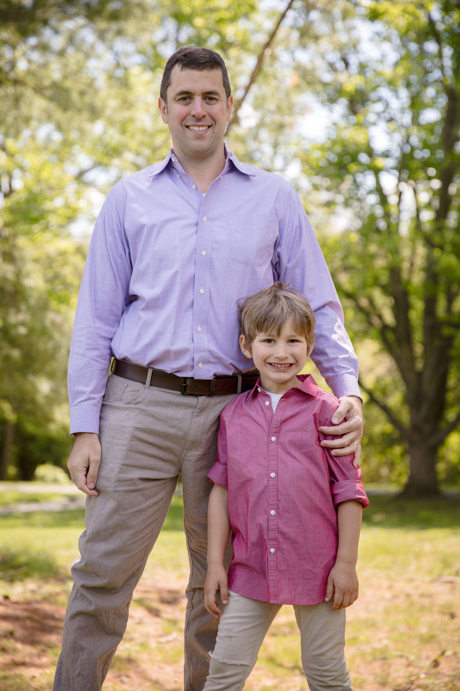 A smiling man in a purple shirt stands with a young boy in a pink shirt outdoors in a sunny park.