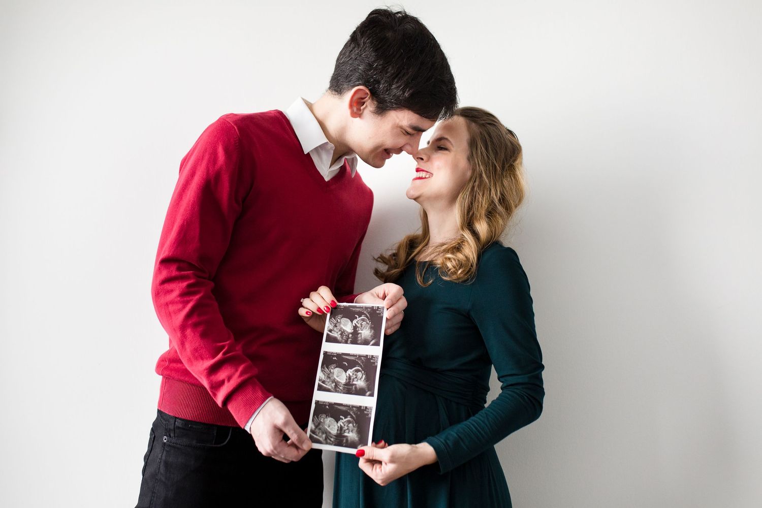 Couple sharing a tender moment while holding ultrasound photos, announcing pregnancy against a white background.