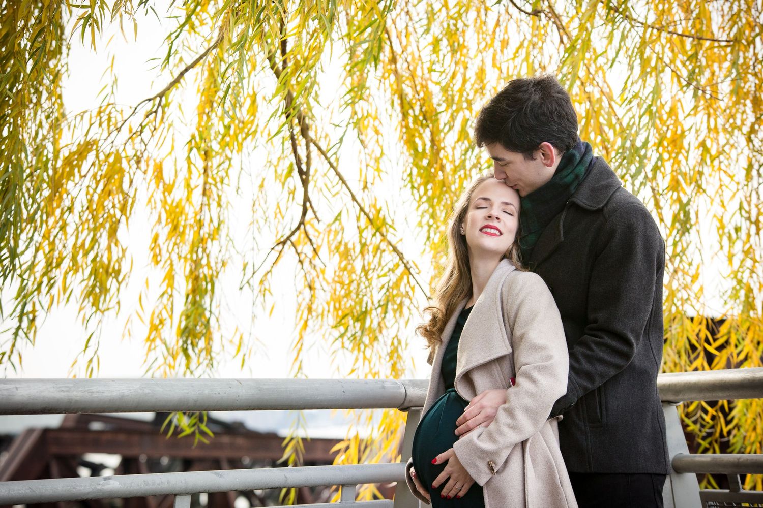 Couple embracing under golden weeping willow tree in autumn, standing on bridge, woman smiling joyfully in beige coat.