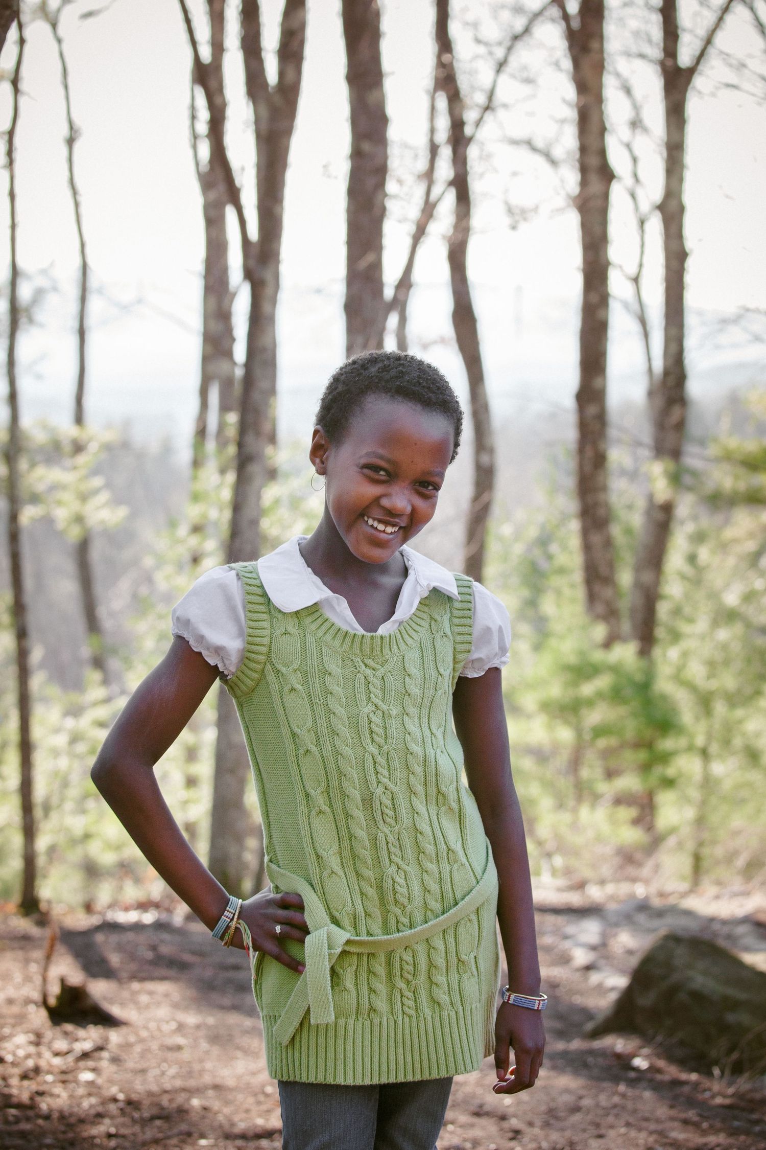 Smiling African girl in green knit vest standing in forest with trees in background.