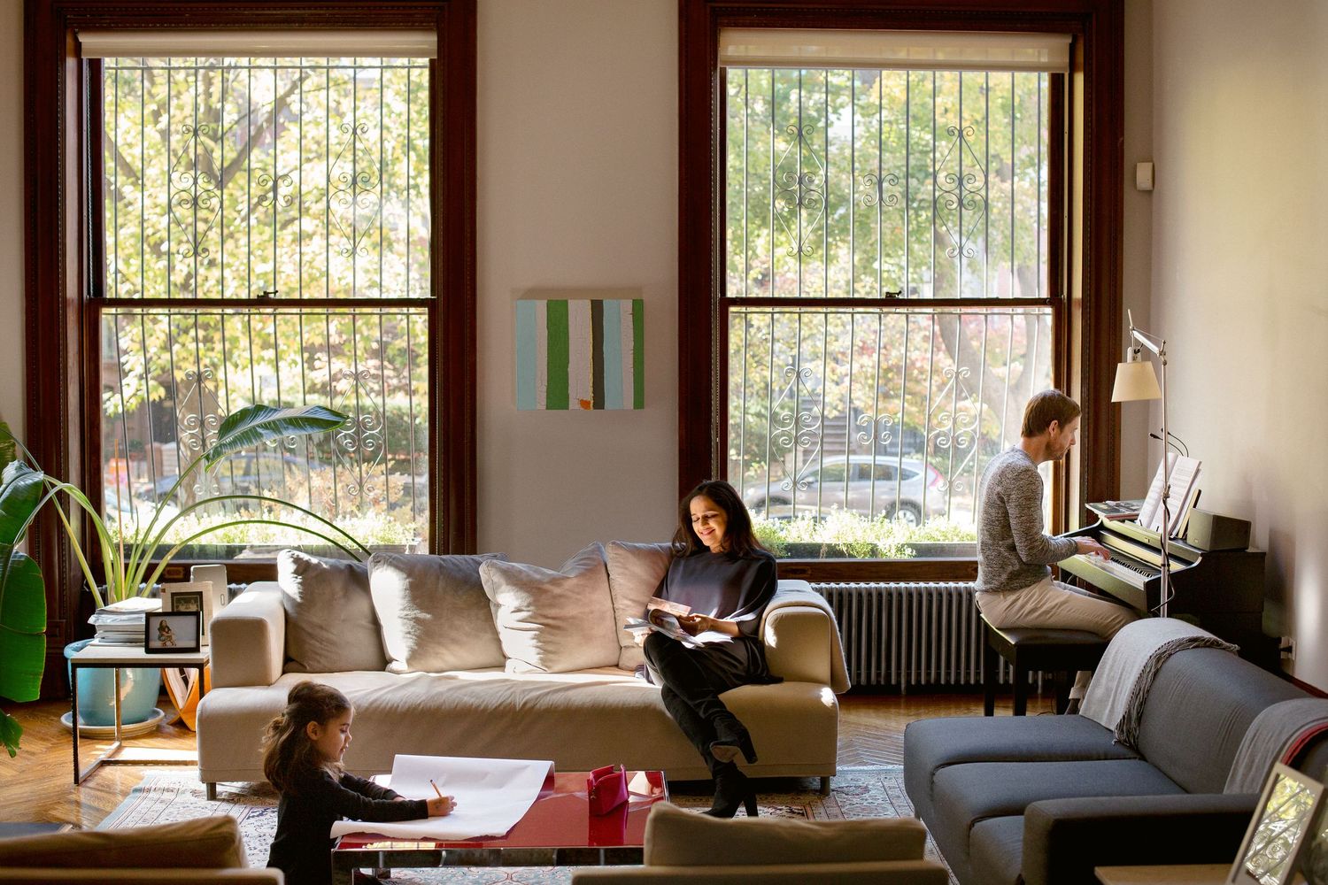 Family relaxing in bright living room with large windows and autumn trees outside.