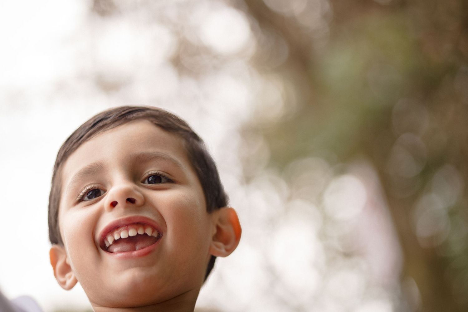 A happy young boy smiling and looking up outdoors with bokeh trees in the background.