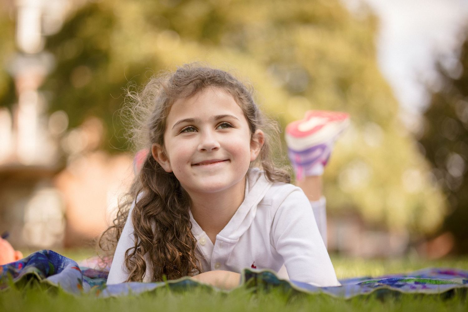 Smiling young girl with curly hair lying on grass in sunny park, looking up cheerfully.